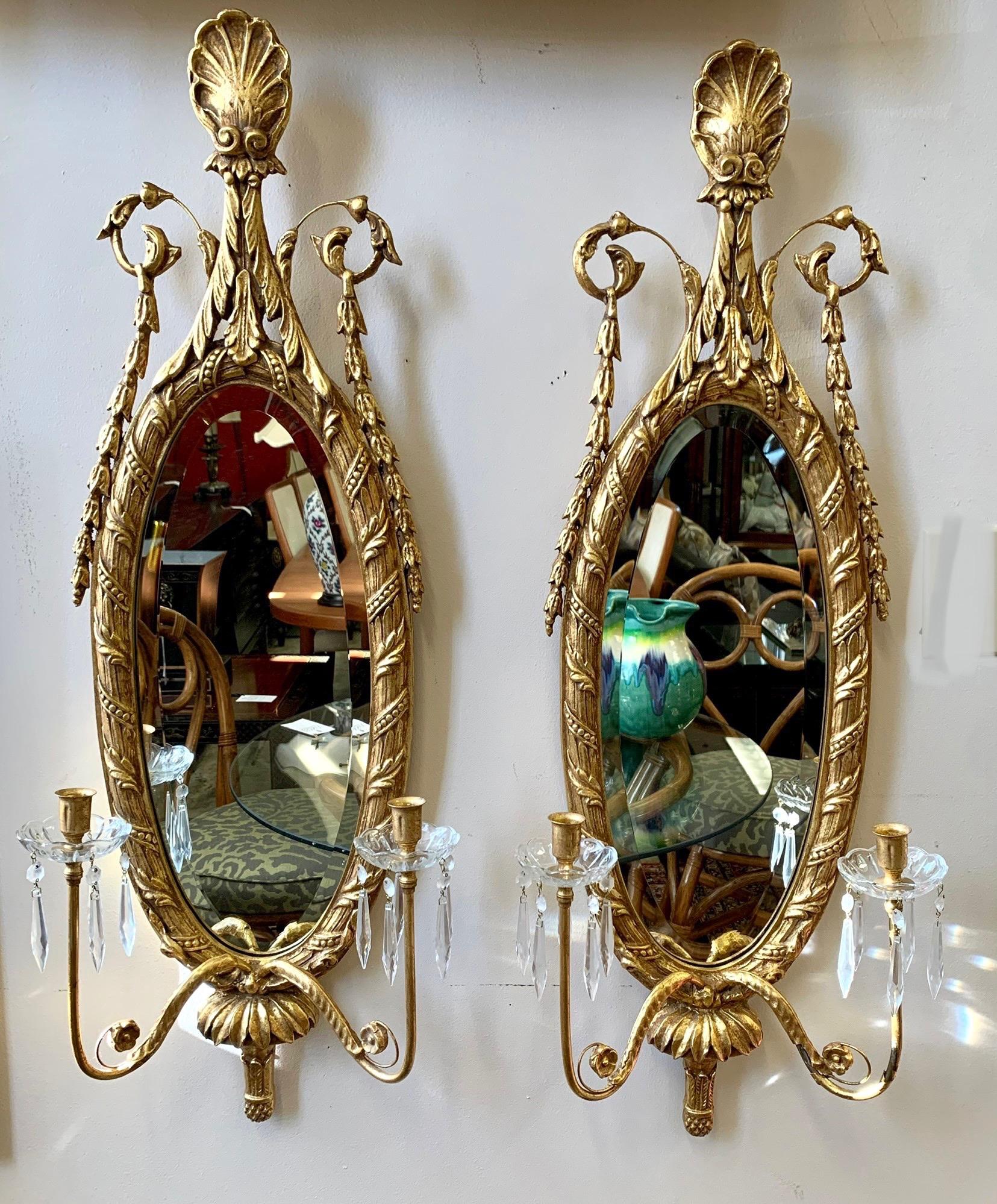Stunning pair of tall carved oval giltwood mirrored sconces with intricate carved details. The two arms with crystal bobeches with dangling crystals hold two candles. Beveled glass. Made in Italy.