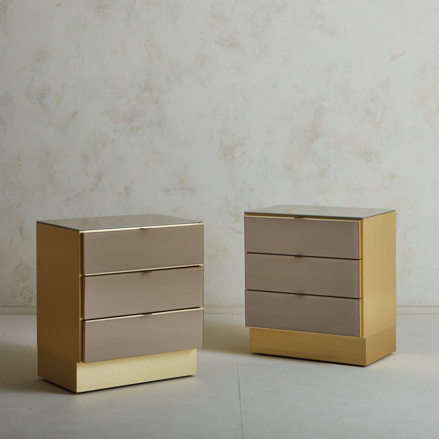 A lovely pair of softly hued back painted glass nightstands by Ello International. These nightstands feature brushed brass along the sides and recessed plinth base, while the back painted glass wraps the three drawer fronts and dresser top. They