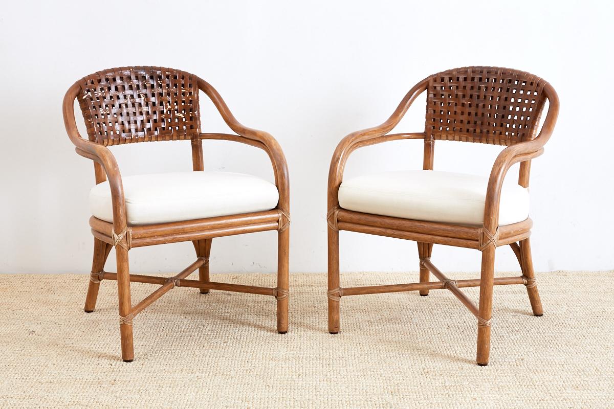 Organic modern pair of genuine McGuire bamboo rattan armchairs featuring a woven leather seat back. Graceful frames constructed from bent rattan with curved arms and seat. Linen upholstered seat cushion and reinforced with leather rawhide strapping.