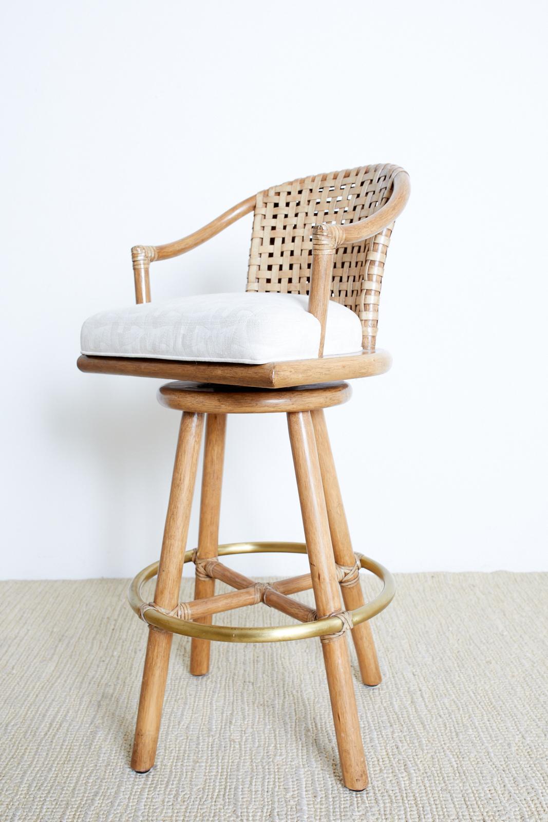 California Modern style pair of swivel barstools by McGuire. Features a bamboo rattan frame and a woven leather backrest. The chairs are reinforced at the joints with leather rawhide laces and have round straight legs conjoined by a brass ring