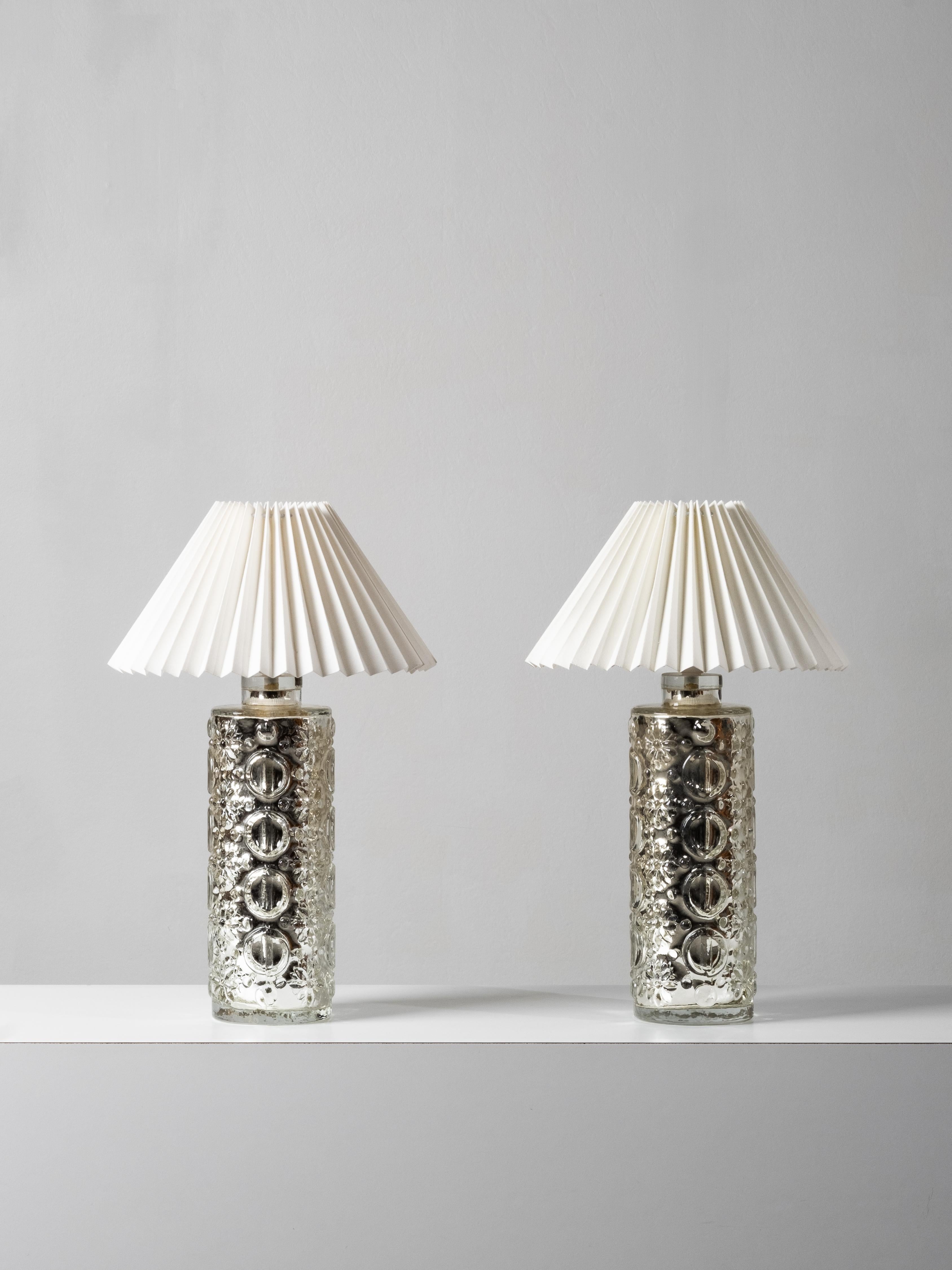 Striking pair of mercury glass table lamps designed by Gustav Leek for Orrefors, Sweden, 1960s. Each lamp features a cylindrical glass body with richly textured relief patterns and circular motifs, finished with a mirrored mercury interior that