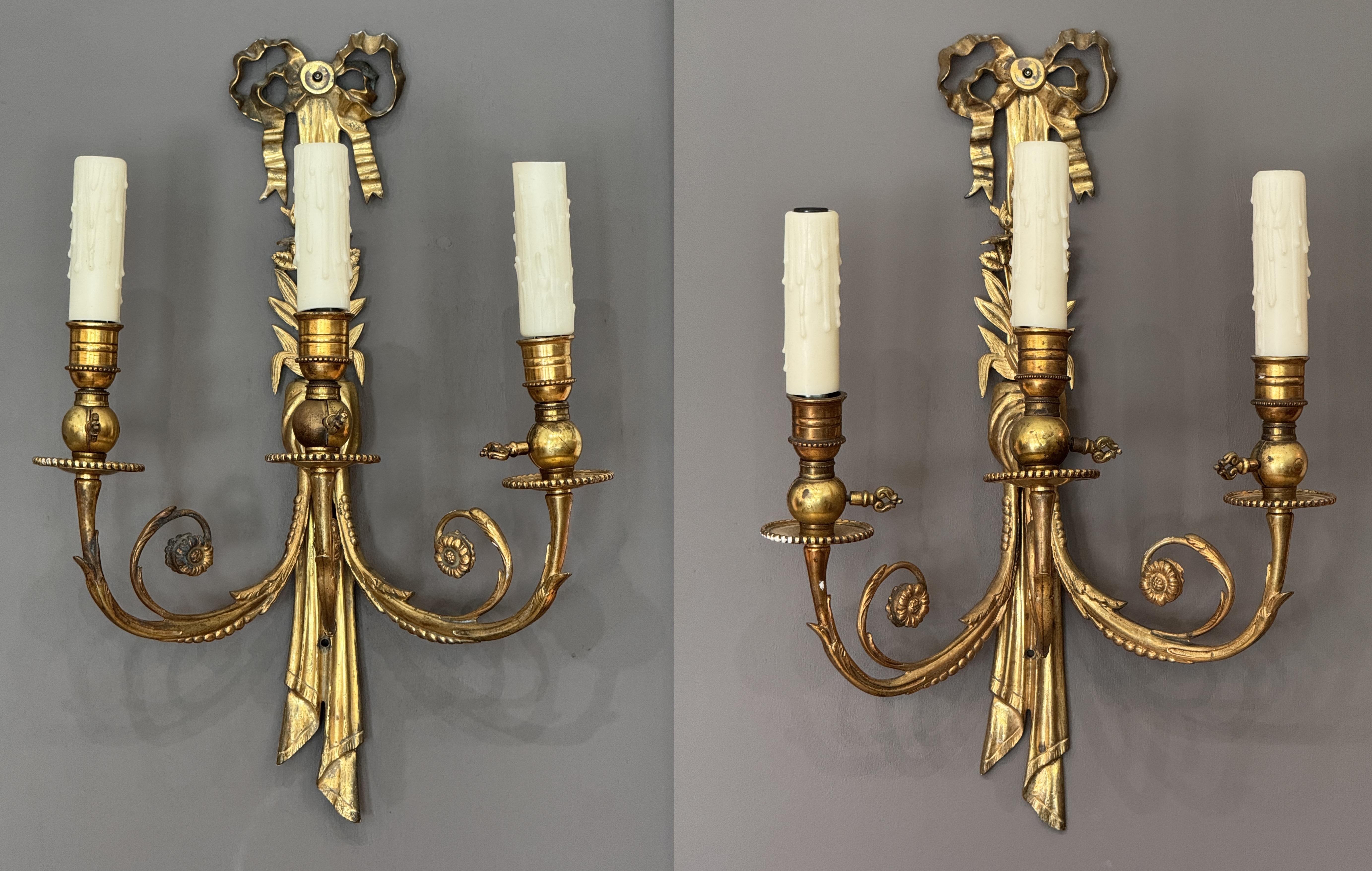 Elegante coppia di appliques francesi in bronzo dorato della metà del XIX secolo, ciascuna con tre bracci a candela e dettagli classici. Il design è caratterizzato da motivi floreali legati a nastri e da una raffinata lavorazione a volute,