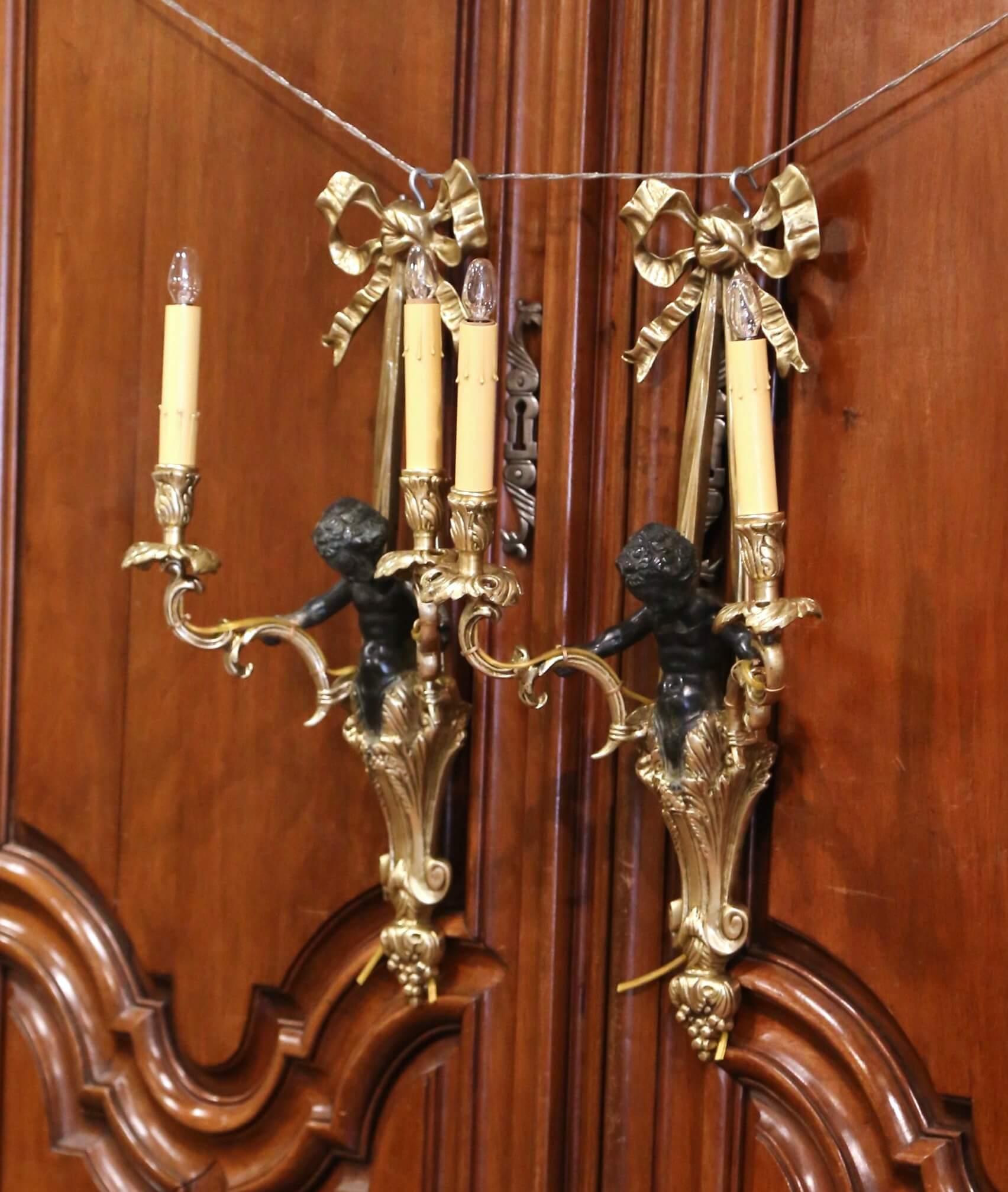 These elegant antique bronze fixtures were crafted in France, circa 1860. Each tall wall sconce features the traditional Louis XVI ribbon bow at the pediment over a patinated cherub figure draped in acanthus leaves and holding the two arms; it has