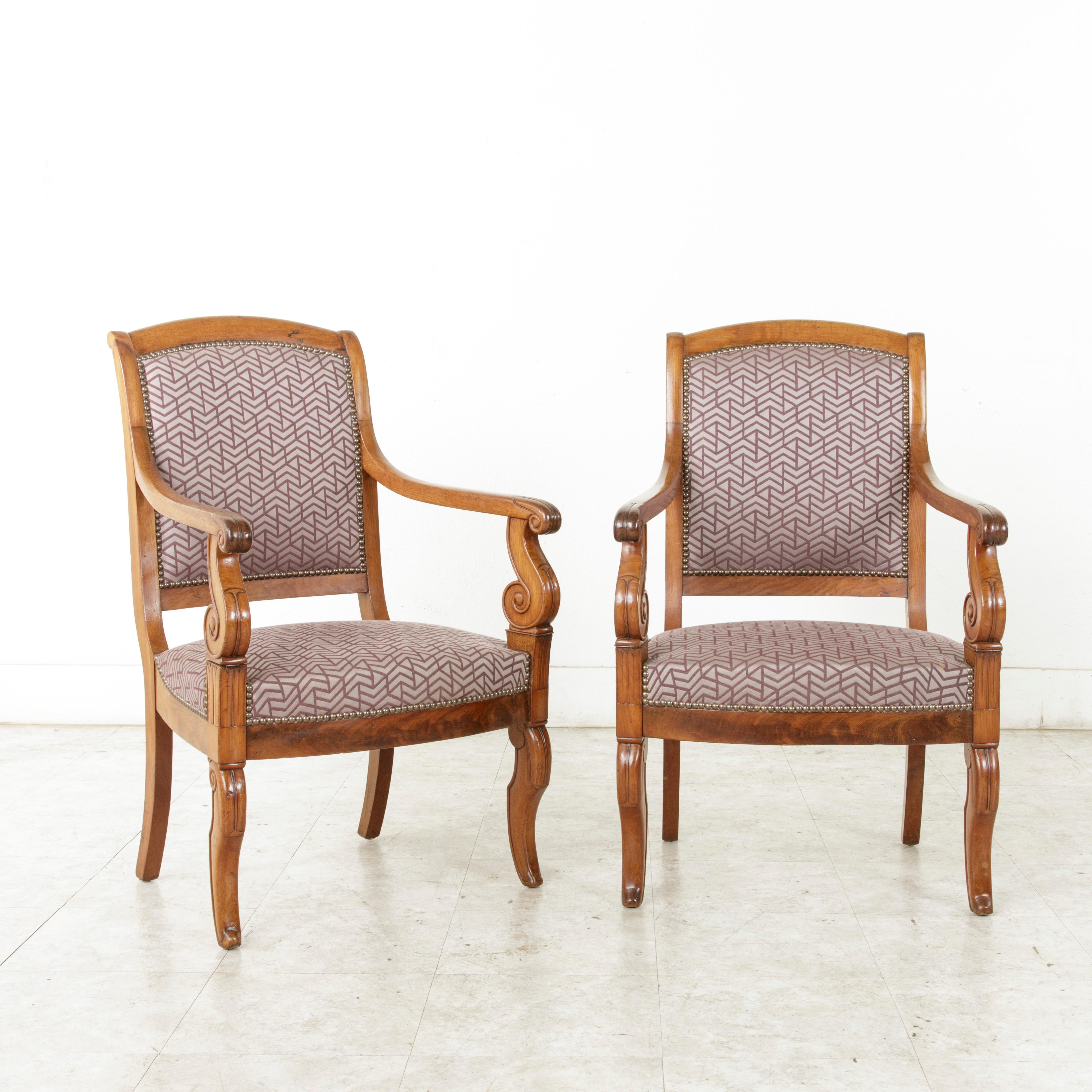 This elegant pair of French Restauration period walnut armchairs from the mid-19th century features beautifully hand carved scrolling arms and Egyptian water leaves on the front die joints. The front curved legs repeat the scrolling of the arms. The