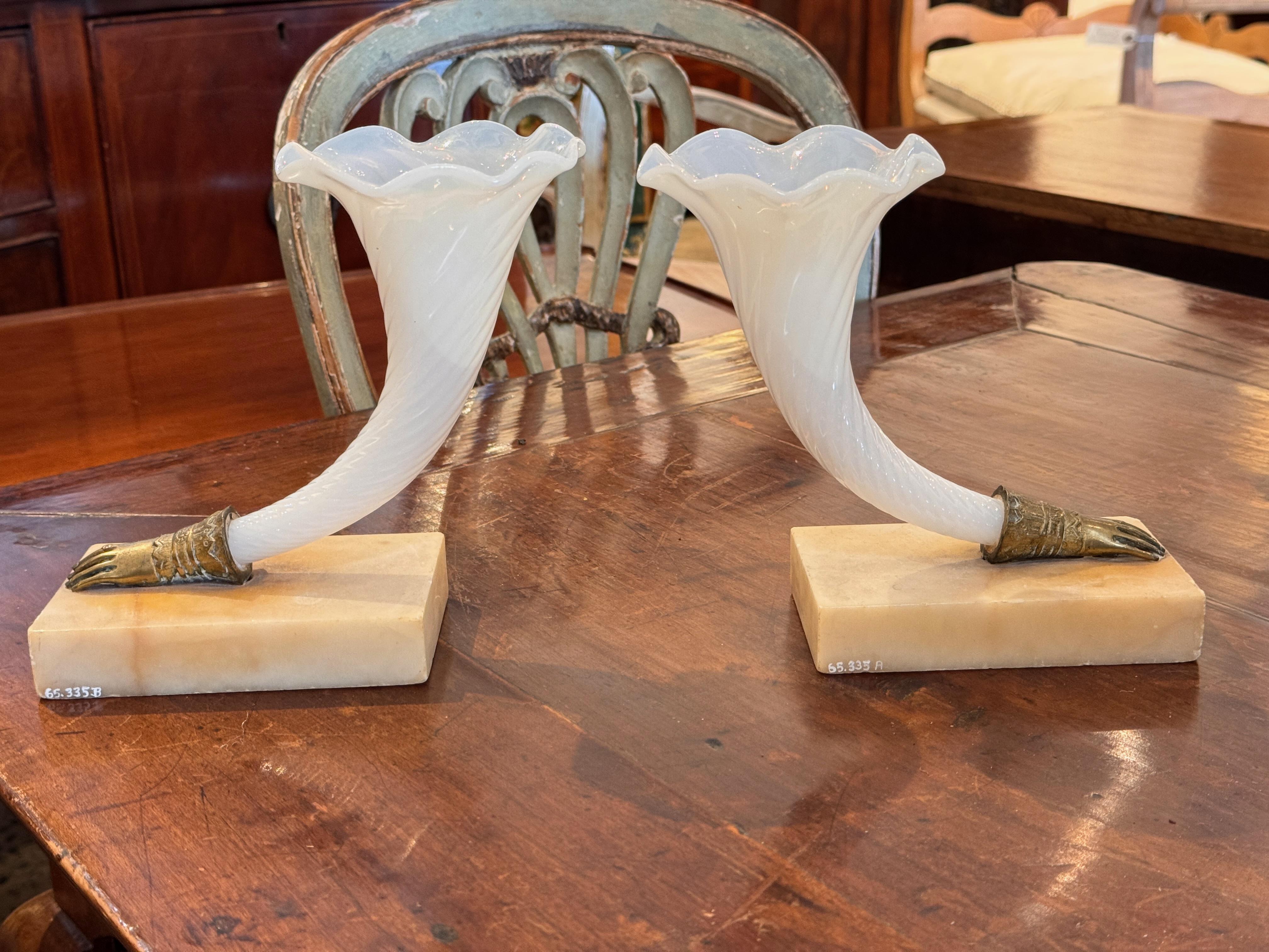 White opaline glass cornucopia vases with twisted bodies and flared rims, each mounted in a cast metal hand-form holder and set on rectangular stone bases. A decorative blend of neoclassical and whimsical detail.