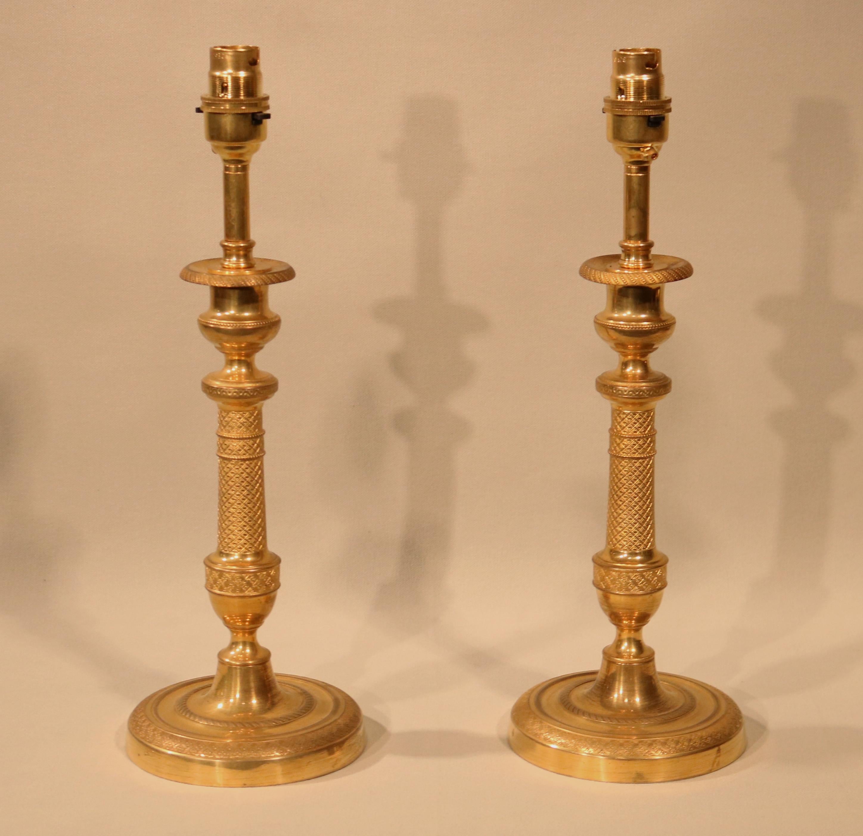 A pair of mid-19th century ormolu candlesticks with engine-turned detail throughout, having urn-shaped nozzles and tapering stems with vase-shape to the bottom, ending on circular bases. (Now converted to lamps.)