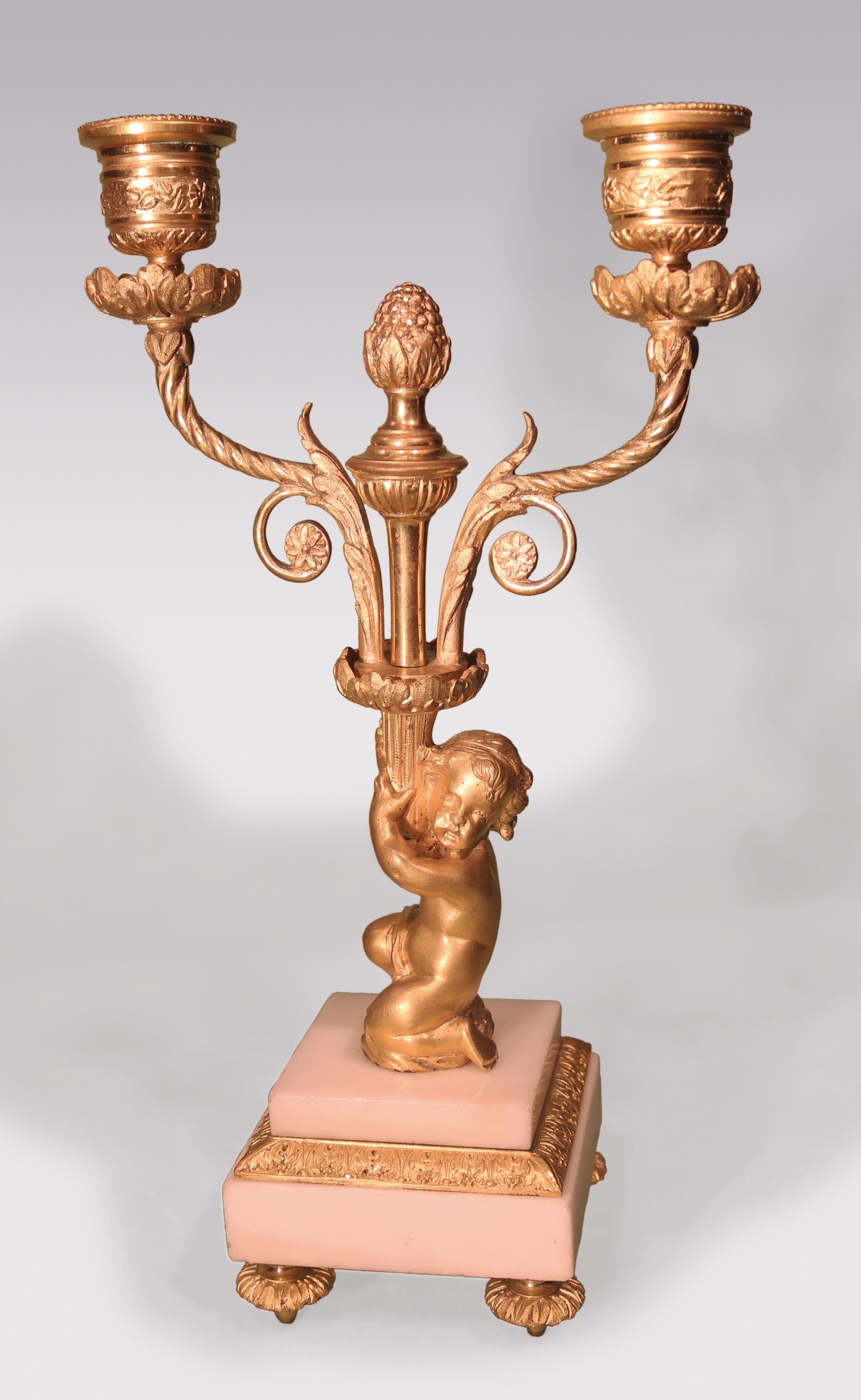 A pair of mid-19th century French ormolu two-light Candelabra, in the form of crouching cherubs holding scrolled candle-arms with central urn and finial, supported on white marble stepped plinth bases, ending on toupe feet.