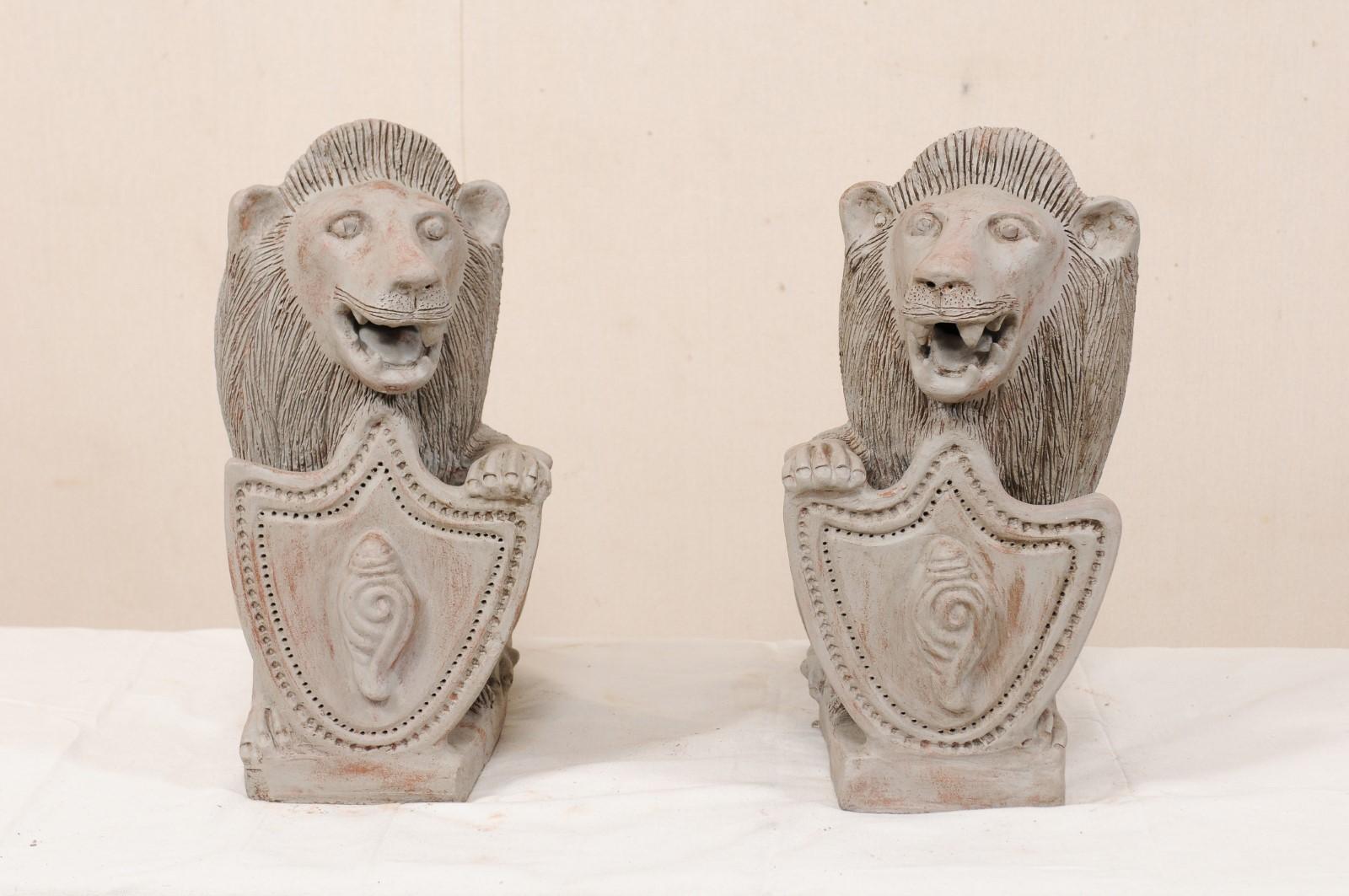 A pair of British Colonial painted terracotta lions from the mid-20th century. This vintage pair of lions from Kerala, India have each been created in a seated position with a single front leg or paw raised atop a shield, which is decorated with a