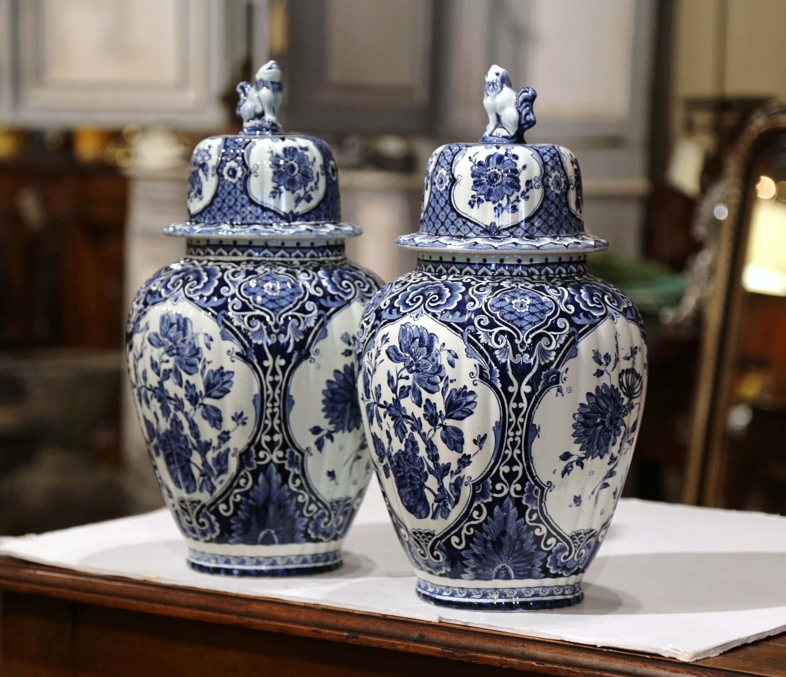 Created in Holland, circa 1960, the faience potiches feature hand painted medallions with Classic Dutch flowers and foliage. Signed delfts, the traditional blue and white jars have a dome lid embellished with dog figure at the top, and are stamped