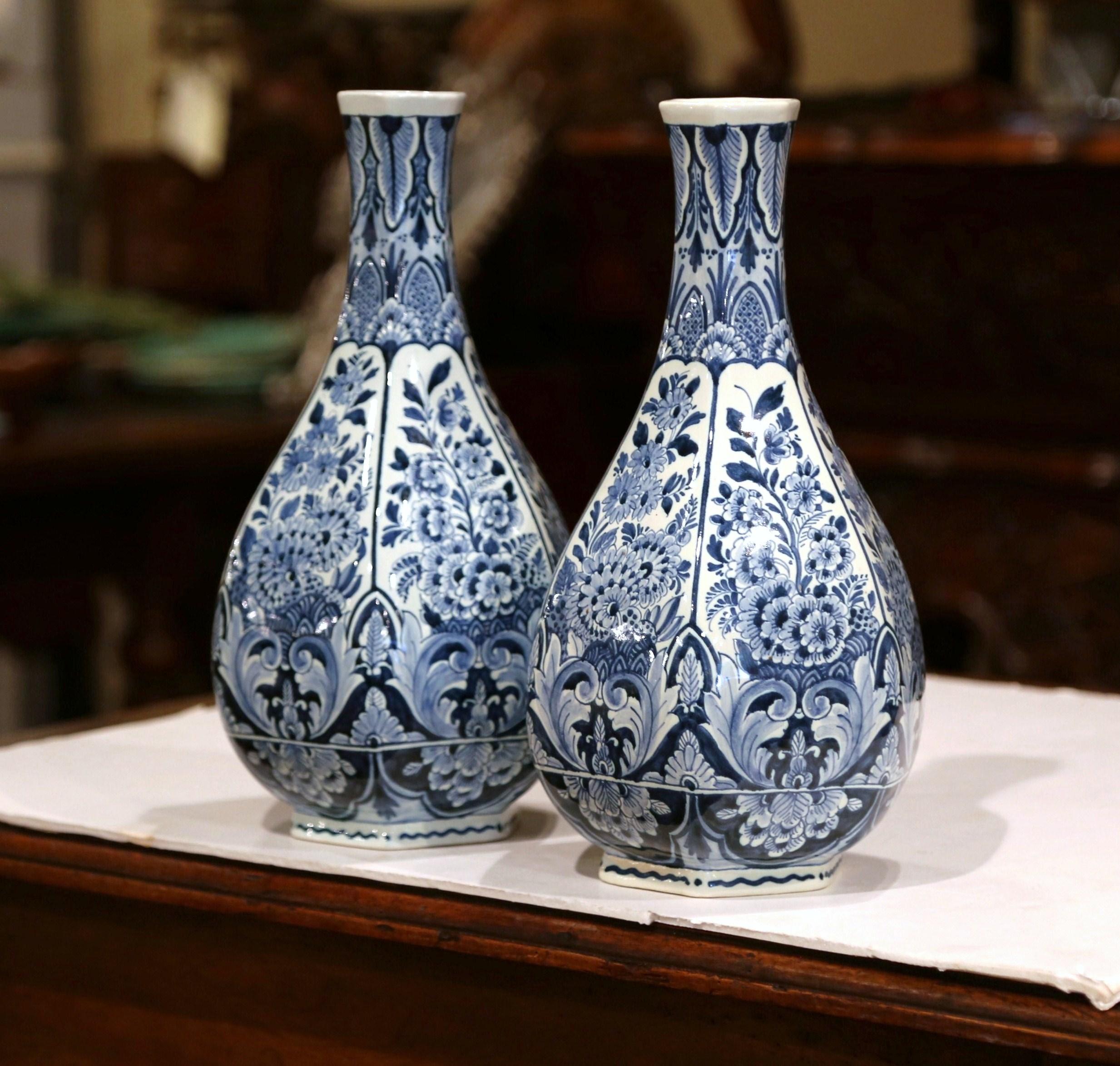 This vintage pair of ceramic delft vases was created in Holland, circa 1950. Hexagonal in shape with a tall and narrow neck, the flower vases feature traditional hand painted floral and leaf decorations in a Classic navy blue and white palette. Both