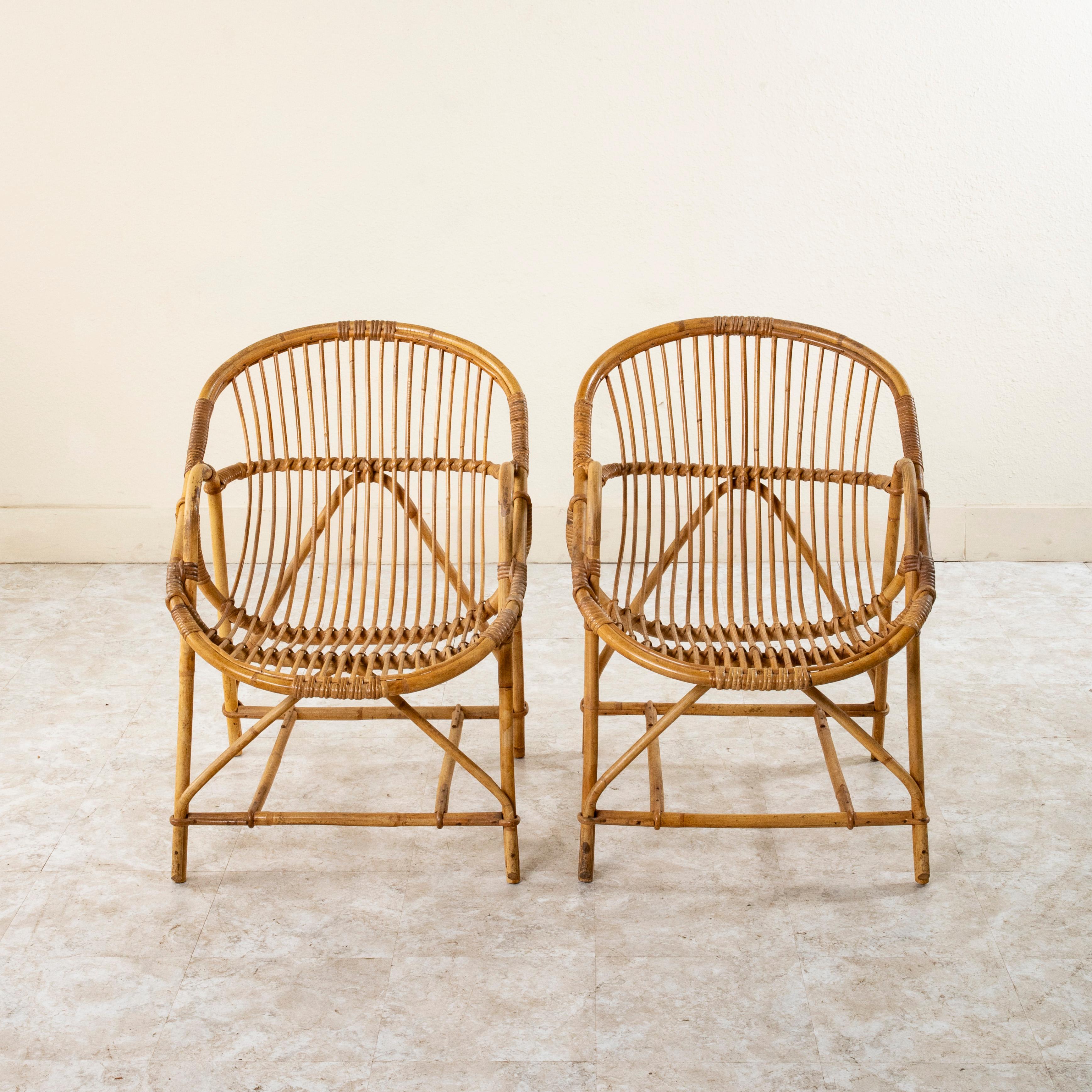 This pair of mid-century French rattan armchairs features bucket shaped seats and backs. The seats rest on curved legs that arch to create armrests. The legs are joined by supports at the front and back and are secured with a double stretcher in