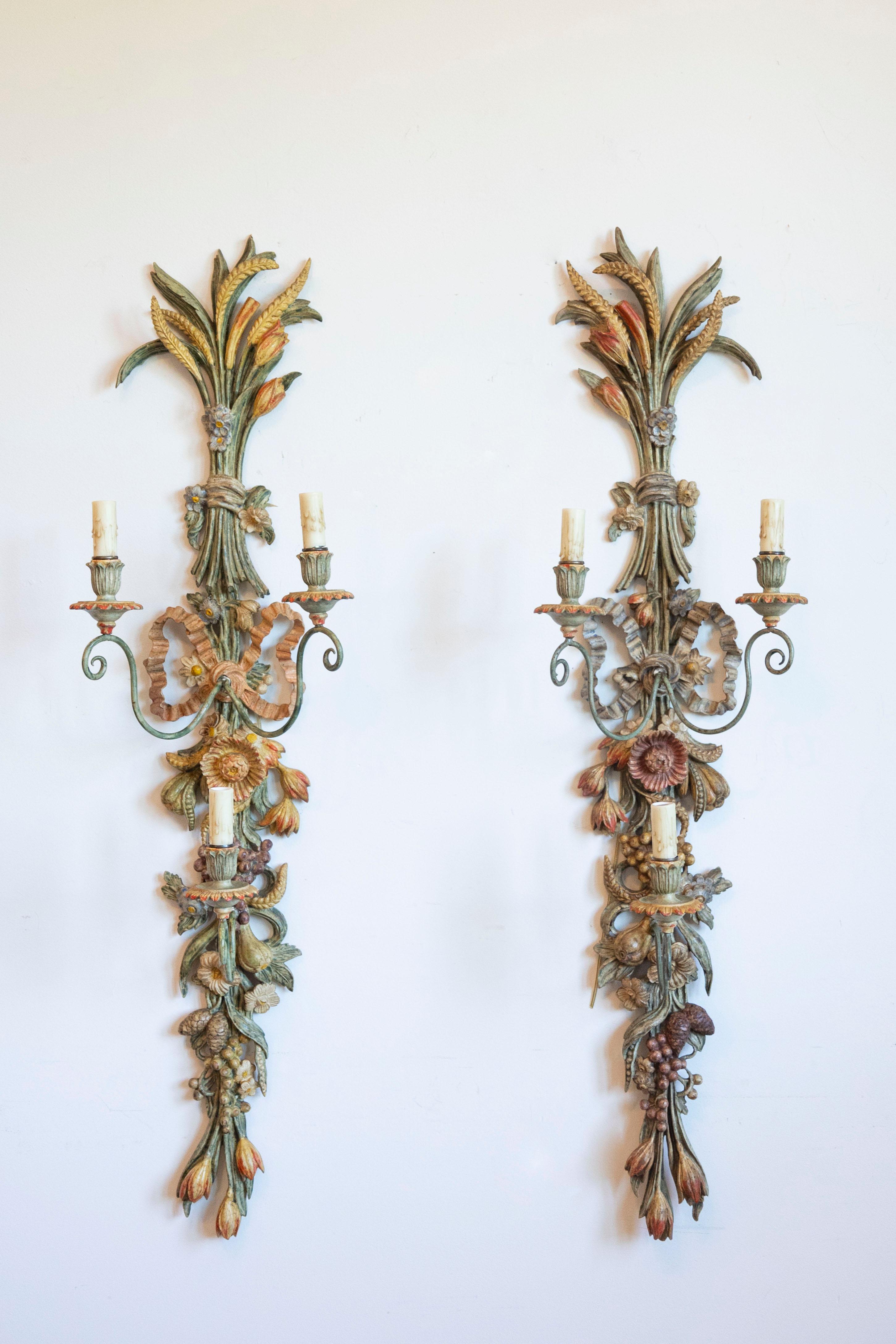 Un'impressionante coppia di applique alte italiane, intagliate a mano nel legno e dipinte a mano in toni morbidi e terrosi. Risalenti alla metà del XX secolo, questi pezzi scultorei sono ornati da covoni di grano, fiori, frutti e fogliame - simboli