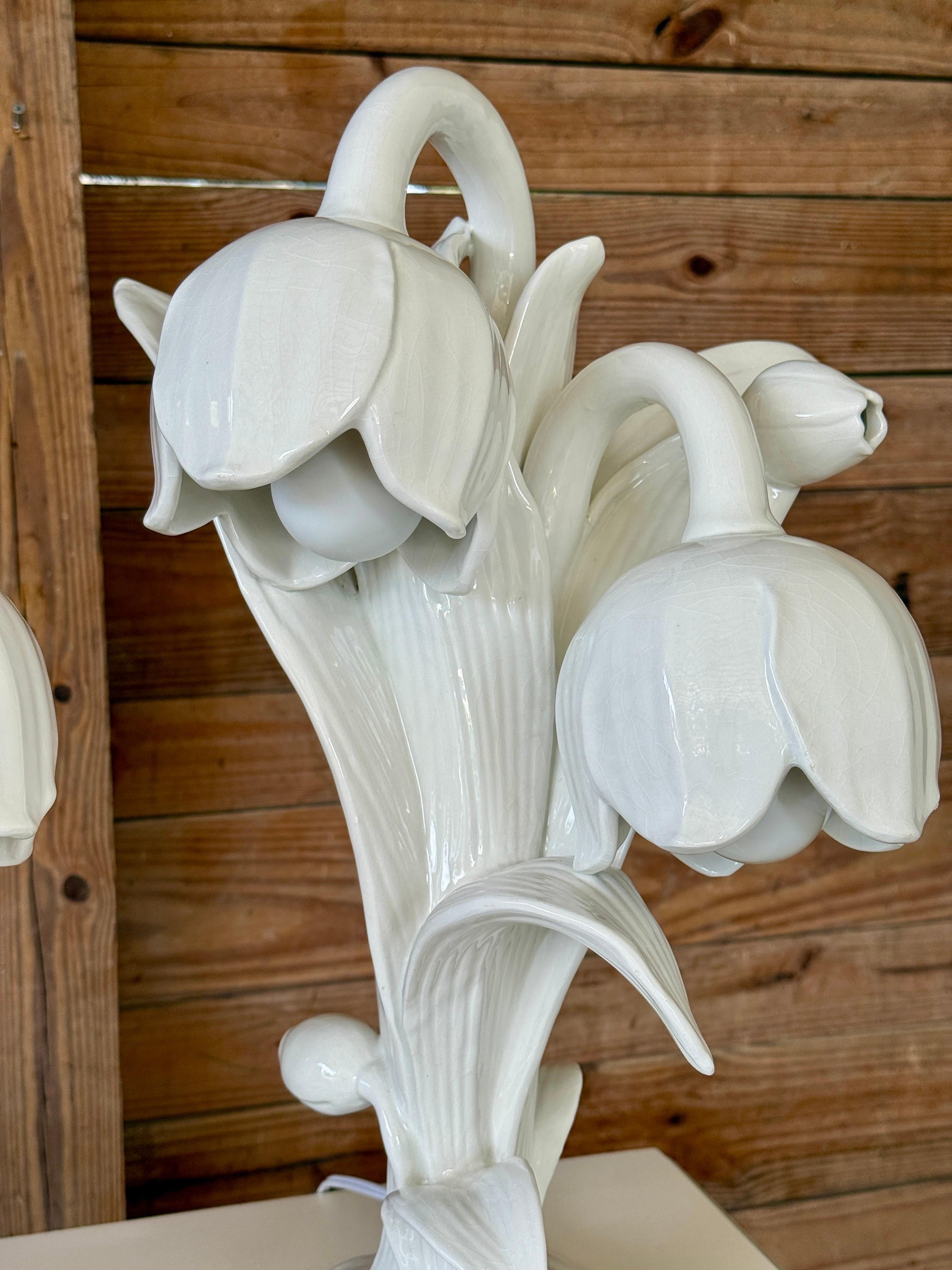 An exquisite and rare pair of Italian porcelain table lamps in the form of sculptural lily blooms, beautifully glazed in a soft white finish. Each lamp is intricately crafted, featuring three upward-curving stems with bell-shaped blossoms that
