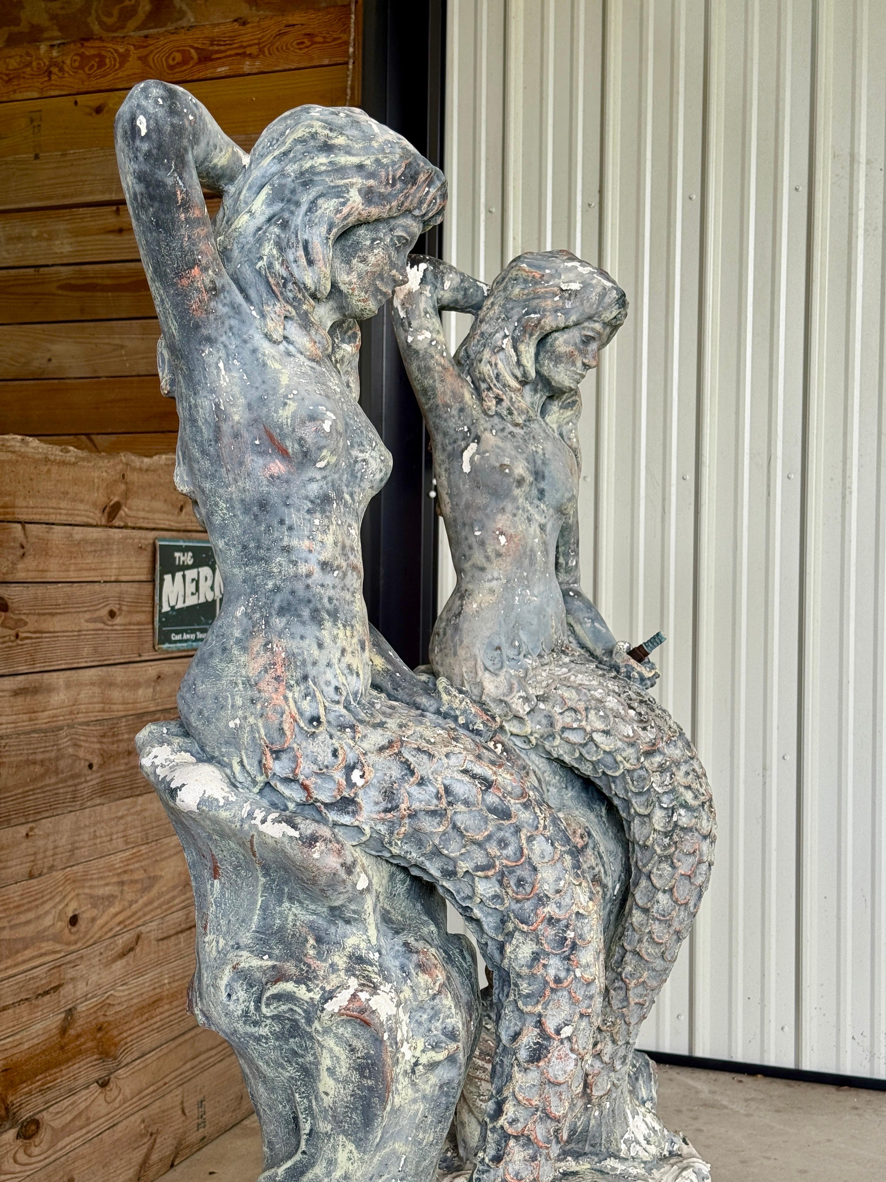 A striking and rare pair of mid-20th century cast concrete mermaid fountain sculptures, each gracefully poised with one arm raised and the other resting near a coiled fish tail. These sculptural figures feature beautifully weathered finishes with