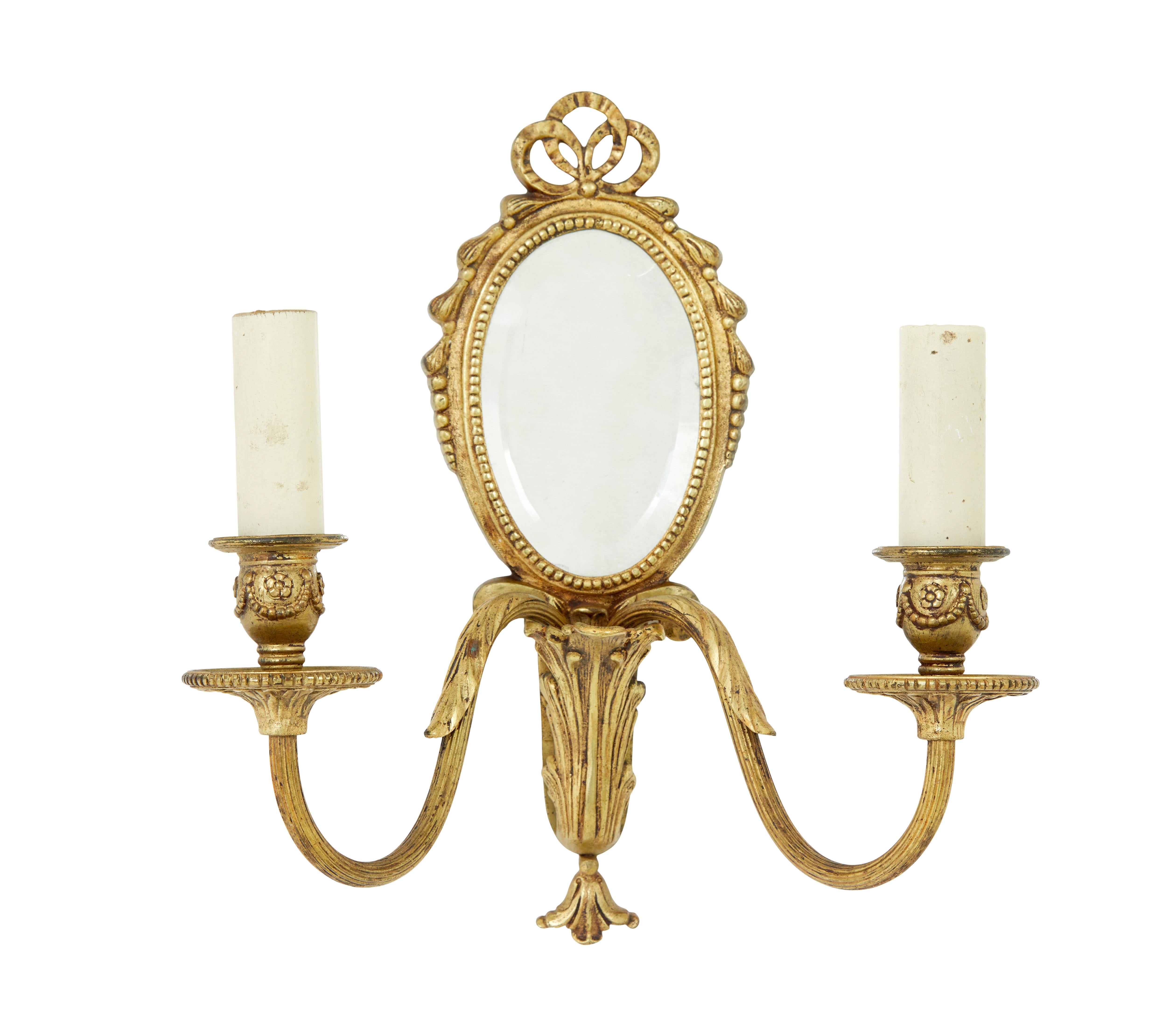 Paar zweiarmige dekorative Wandleuchten um 1960.

Ovaler Spiegel mit abgeschrägter Kante, umgeben von Perlenbordüren und Bügeln.  Geformte Arme mit einer Leuchte, die aus einer Urne herausragt. Originale, gefälschte Kerzenabdeckungen.

Einige