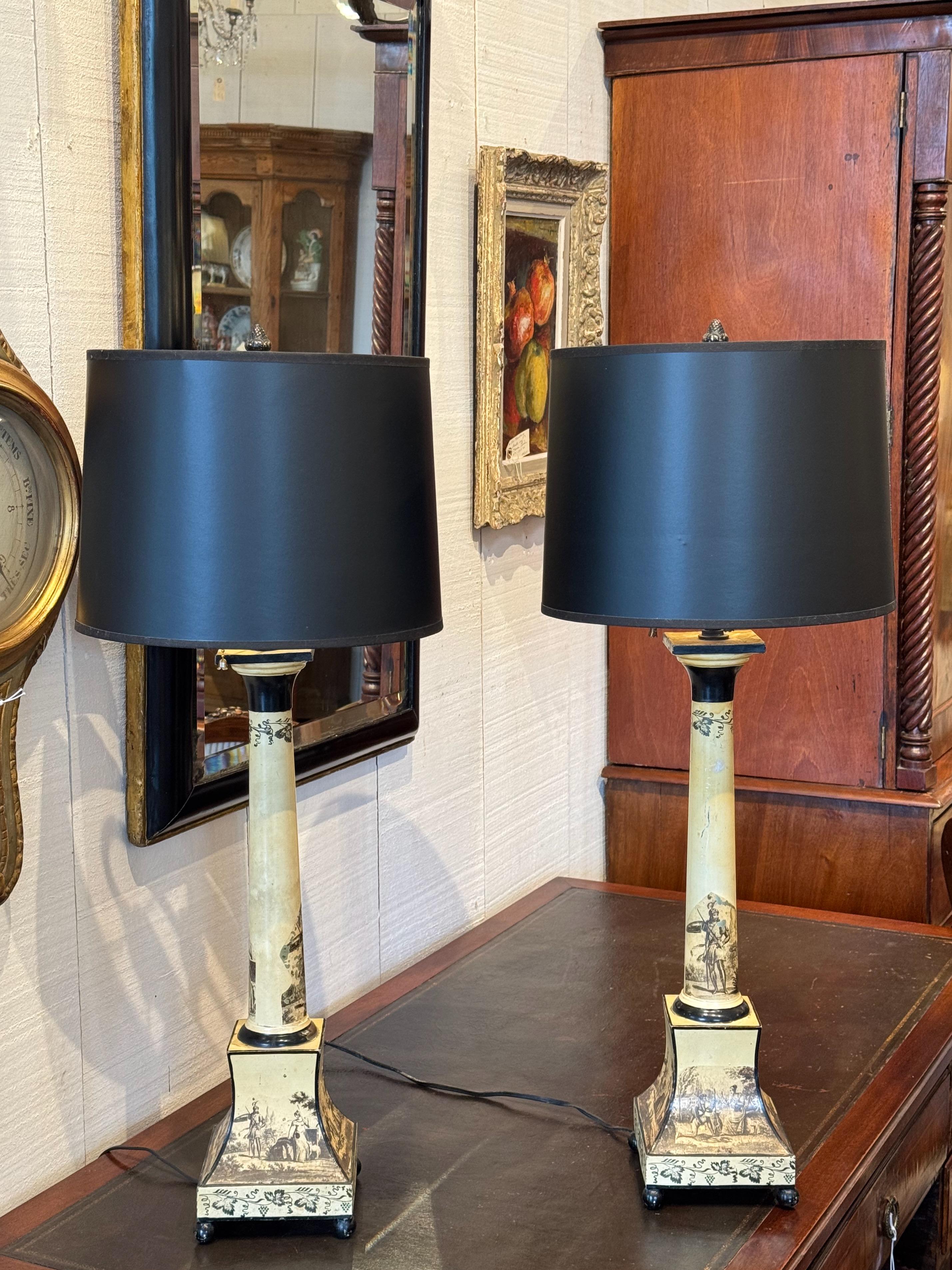 Paire de lampes à tole du milieu du 20e siècle, avec des colonnes effilées sur des bases en forme de socle, peintes à la main dans un motif pastoral classique. La palette noire et crème est accentuée par des fleurs de vigne et des scènes figuratives