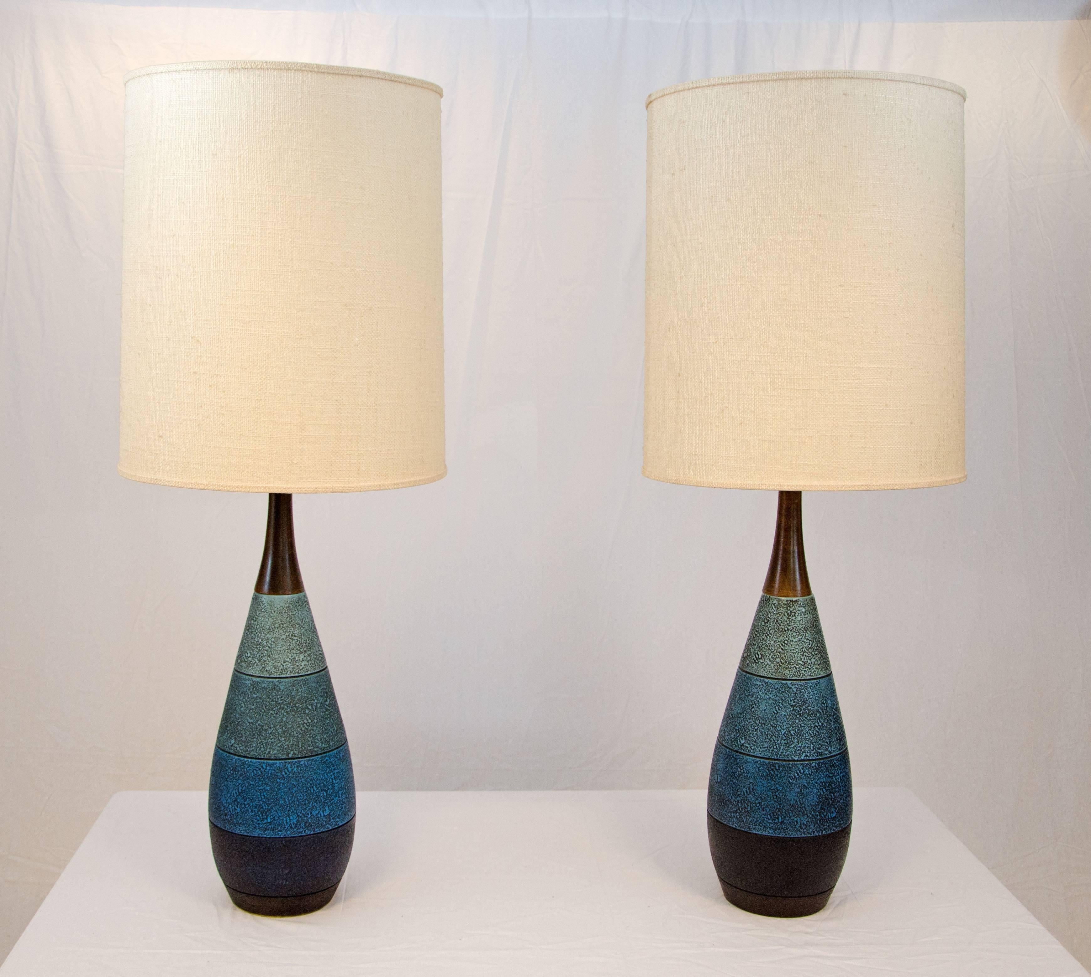 A nice pair of ceramic lamps with original textured fabric shades (thin plastic backing). The ceramic bases are designed with graduated shades of blue and accented with a walnut finished wood. The blue ceramic part of the base is 18