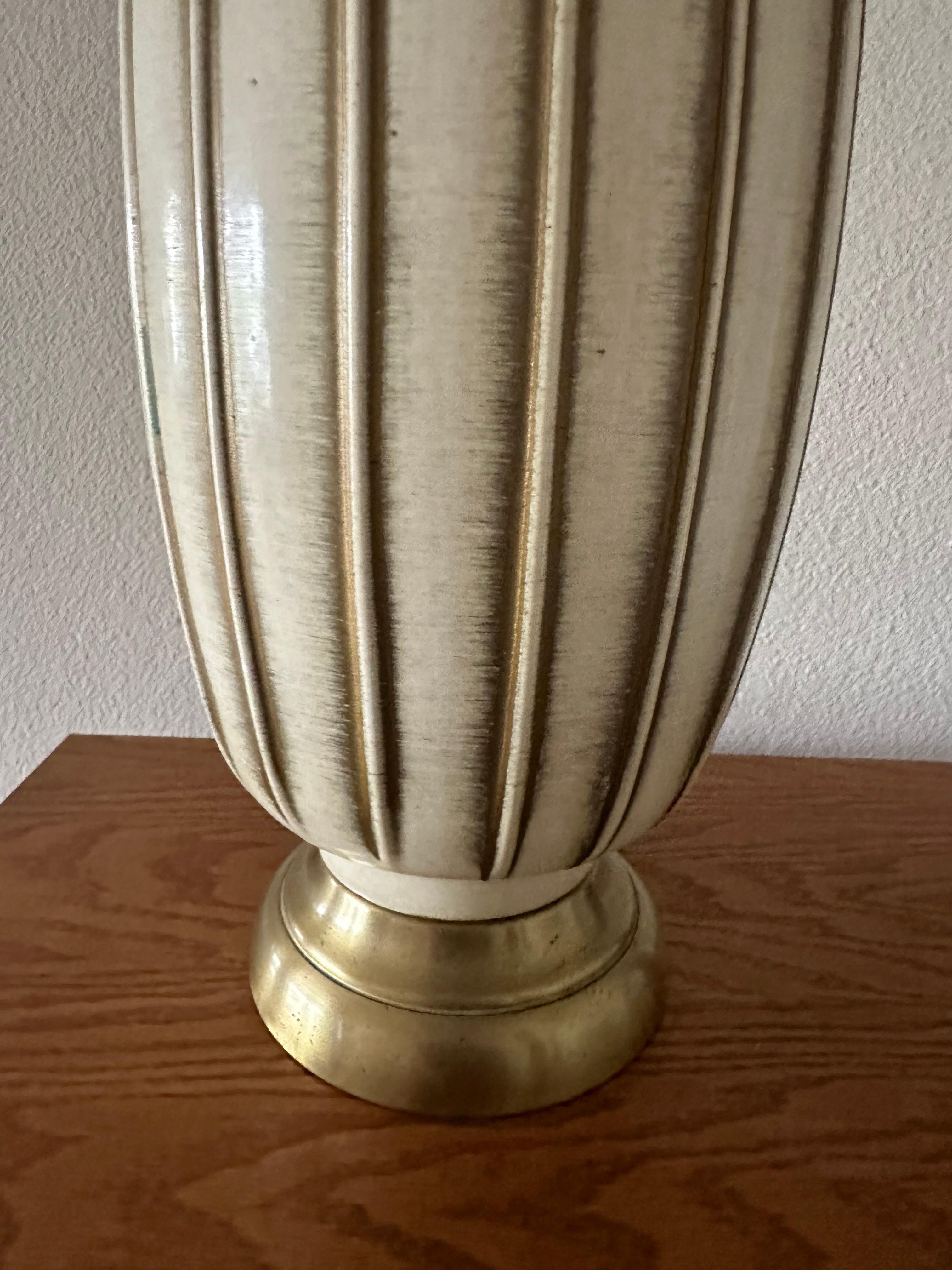 This pair of Mid-Century table lamps features ceramic bodies finished in a soft cream tone with vertically fluted detailing highlighted by subtle golden accents. The rounded forms rest on brushed brass-tone metal bases, adding a warm contrast to the