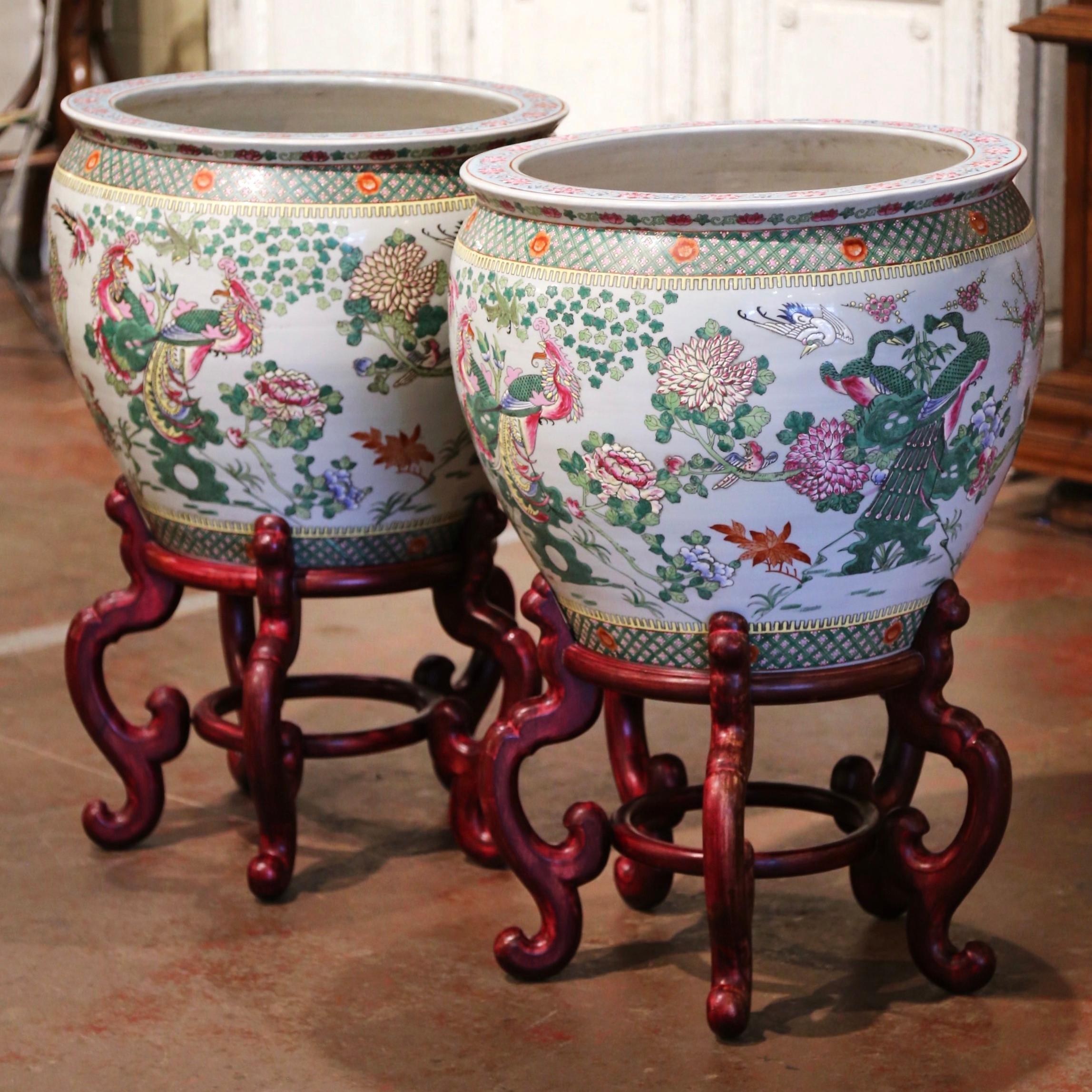These elegant and colorful vintage fishbowls standing on carved fruitwood bases were created in China, circa 1970. Round in shape, each large exotic porcelain planter features Classic bird and peacock motifs embellished with floral decor throughout.