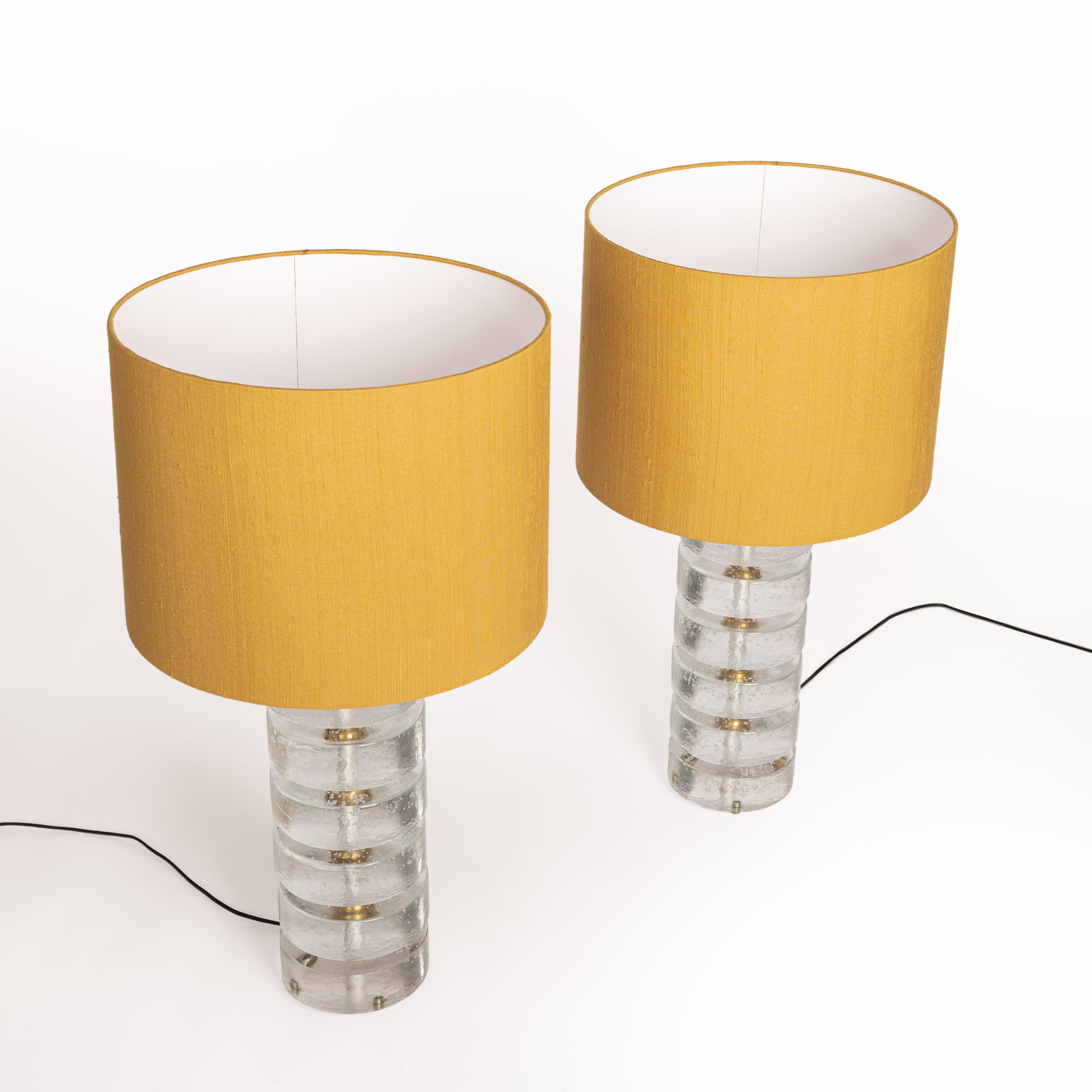 Pair of cylindrical shaped Murano glass table lamps with brass mounting.
The feet are built up of several round glass slices (diameter 16cm x height 5cm) 
and brass supports.
Hand-manufactured lamp shades in mustard-yellow bourette silk and