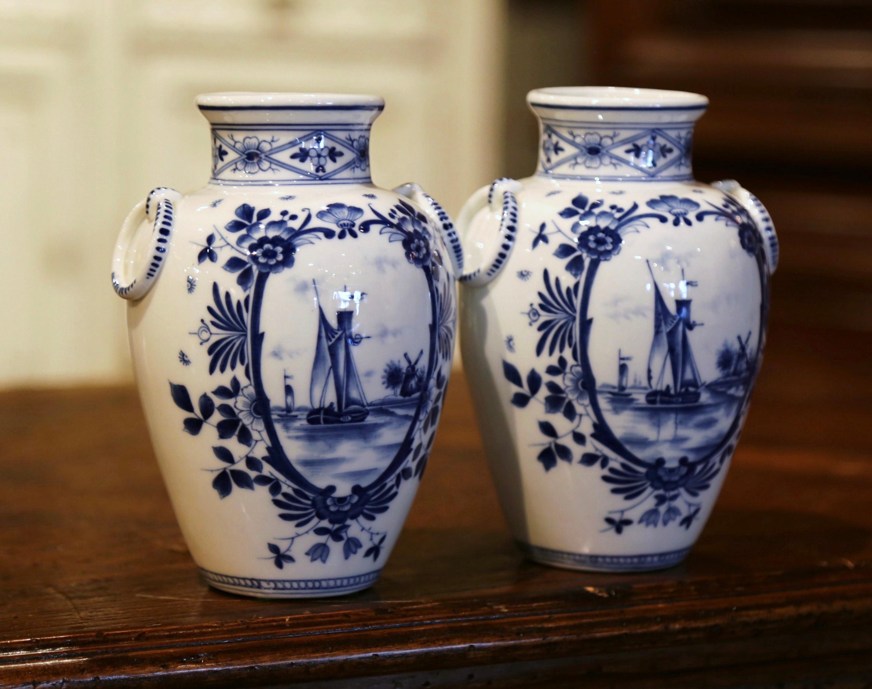 Decorate a shelf or console table with this elegant and colorful pair of antique Delft vases. Crafted in Holland circa 1950, each flower holder is round in shape decorated with small side handle; the vessel is hand painted in the traditional blue