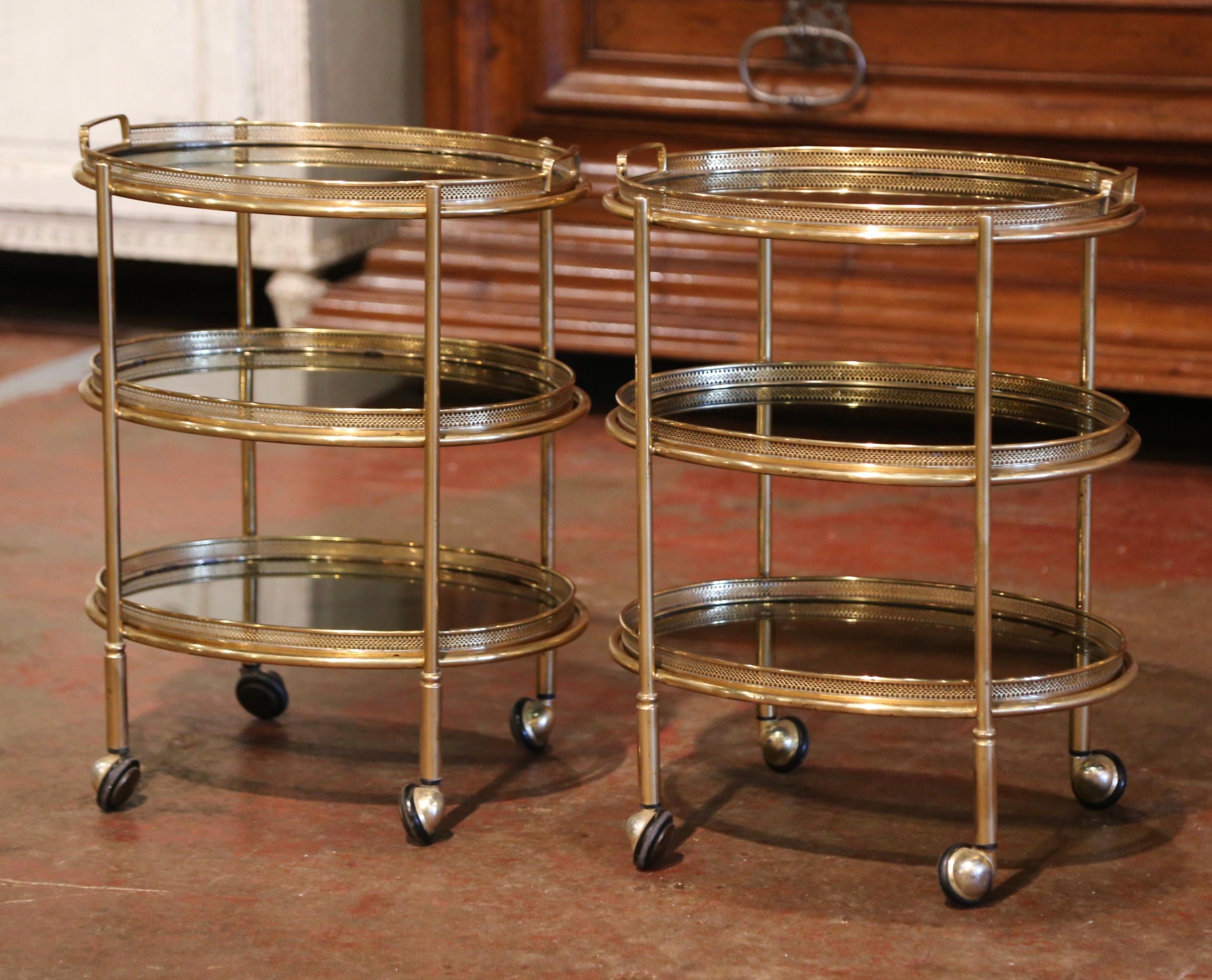 These elegant vintage trolleys bar carts on casters, were created in France, circa 1950. Built in brass and oval in shape, each table stands on small round wheels, over three removable plateaus topped with smoked glass surfaces. The top deck is