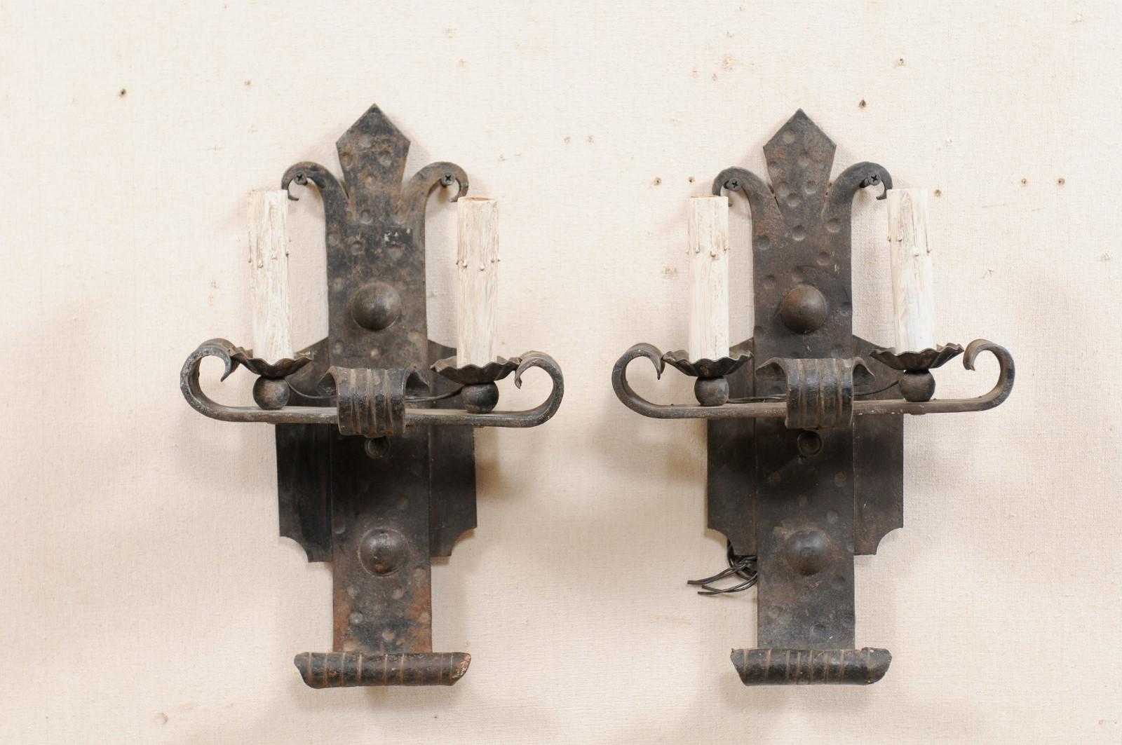 A pair of French two-light sconces from the mid-20th century. These vintage French hand-forged sconces each feature a horizontally placed bar, with upwardly scrolled ends, providing support to the two ruffled iron bobeches with painted candle
