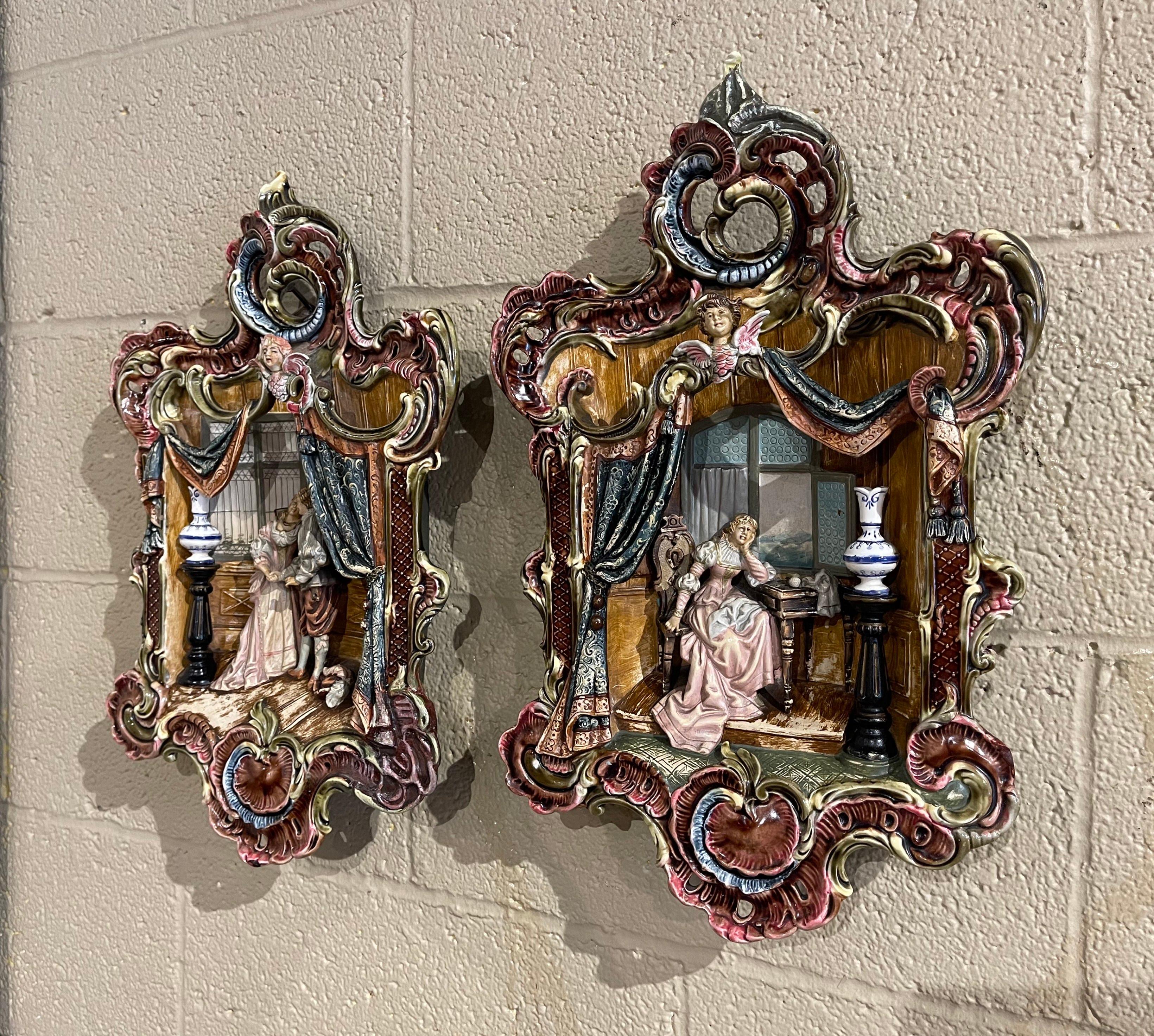 Embellish a wall or shelf with this elegant pair of antique majolica wall hanging plaques. Crafted in France, circa 1960, each ceramic artwork depict a inside interior with man and a woman dressed in traditional Renaissance clothing. Both figures