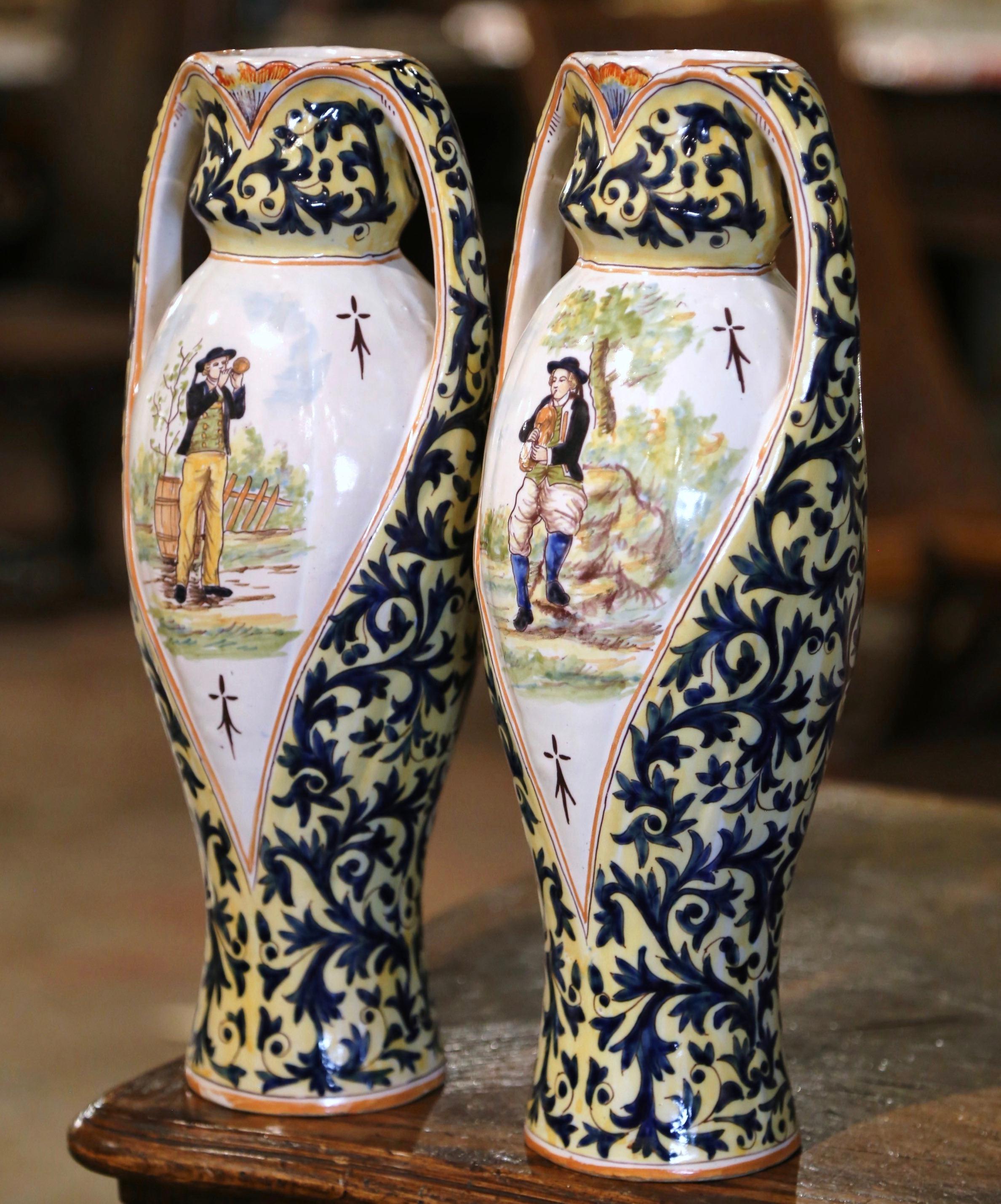 These elegant antique vases were crafted in Brittany France, circa 1940. The tall circular ceramic vases are dressed with side handles over a round neck. Each colorful vessel features hand painted Breton people in traditional clothing, embellished