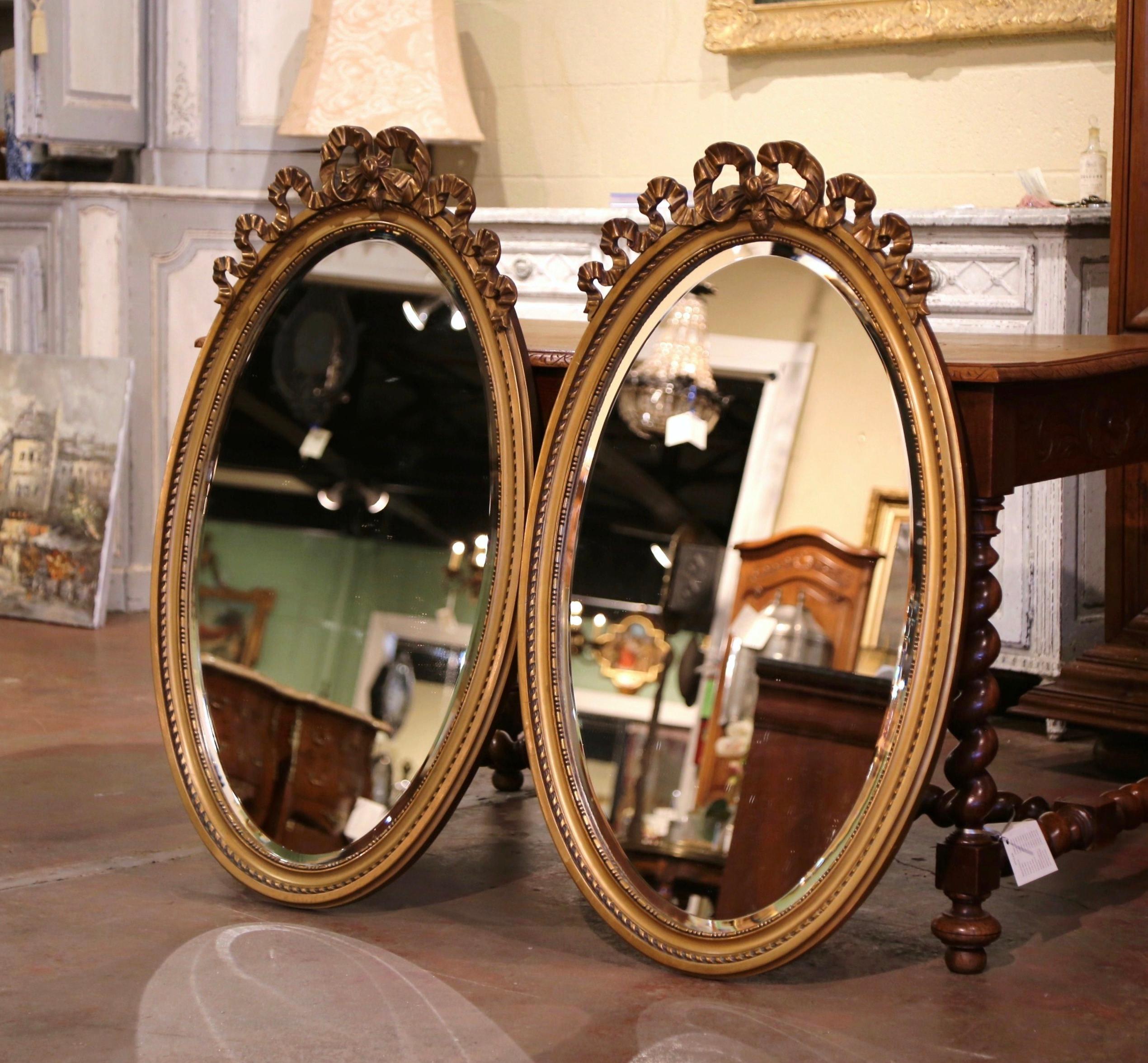 Place these elegant antique mirrors over a pair of consoles or chests! Crafted in France circa 1950 and oval in shape, each wall mirror dressed a beveled glass, features the traditional carved Louis XVI ribbon bow at the pediment embellished with