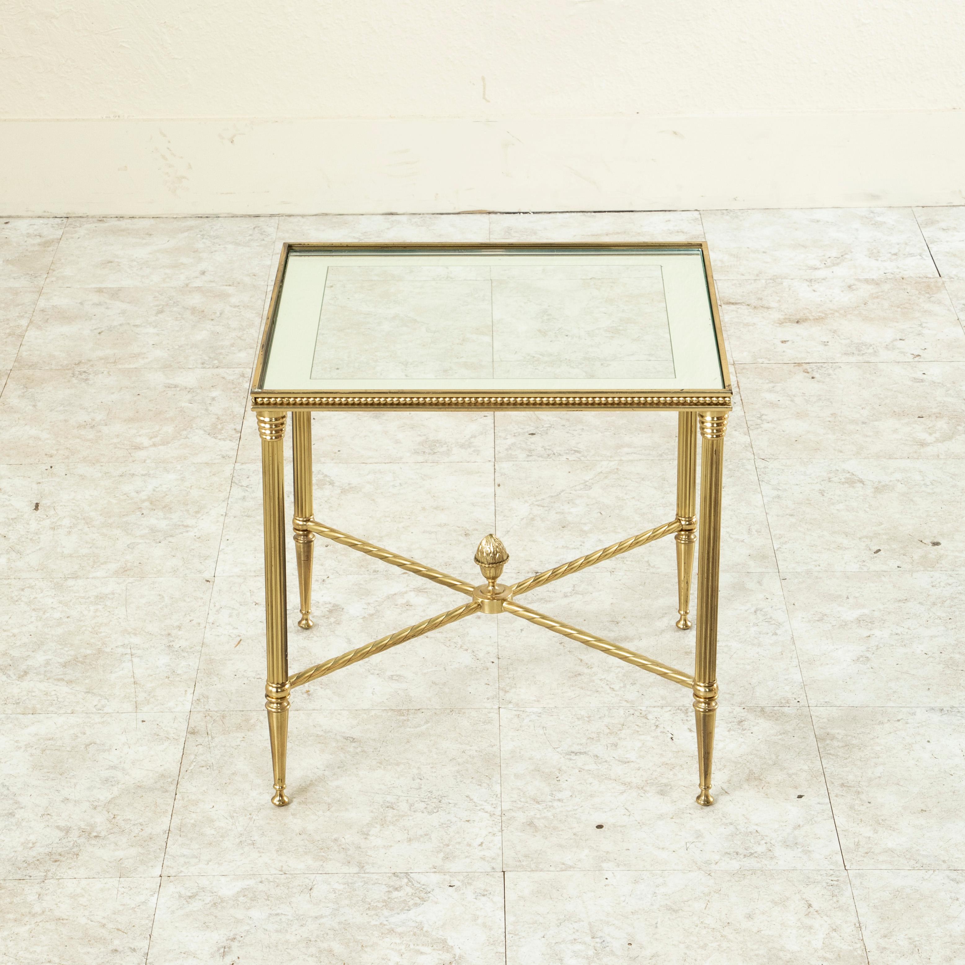 This pair of mid-century Maison Bagues brass side tables features glass tops with mirrored edges. The tables are detailed with fluted legs, beading, and a lower X stretcher with a central Louis XVI acorn finial. c. 1960.