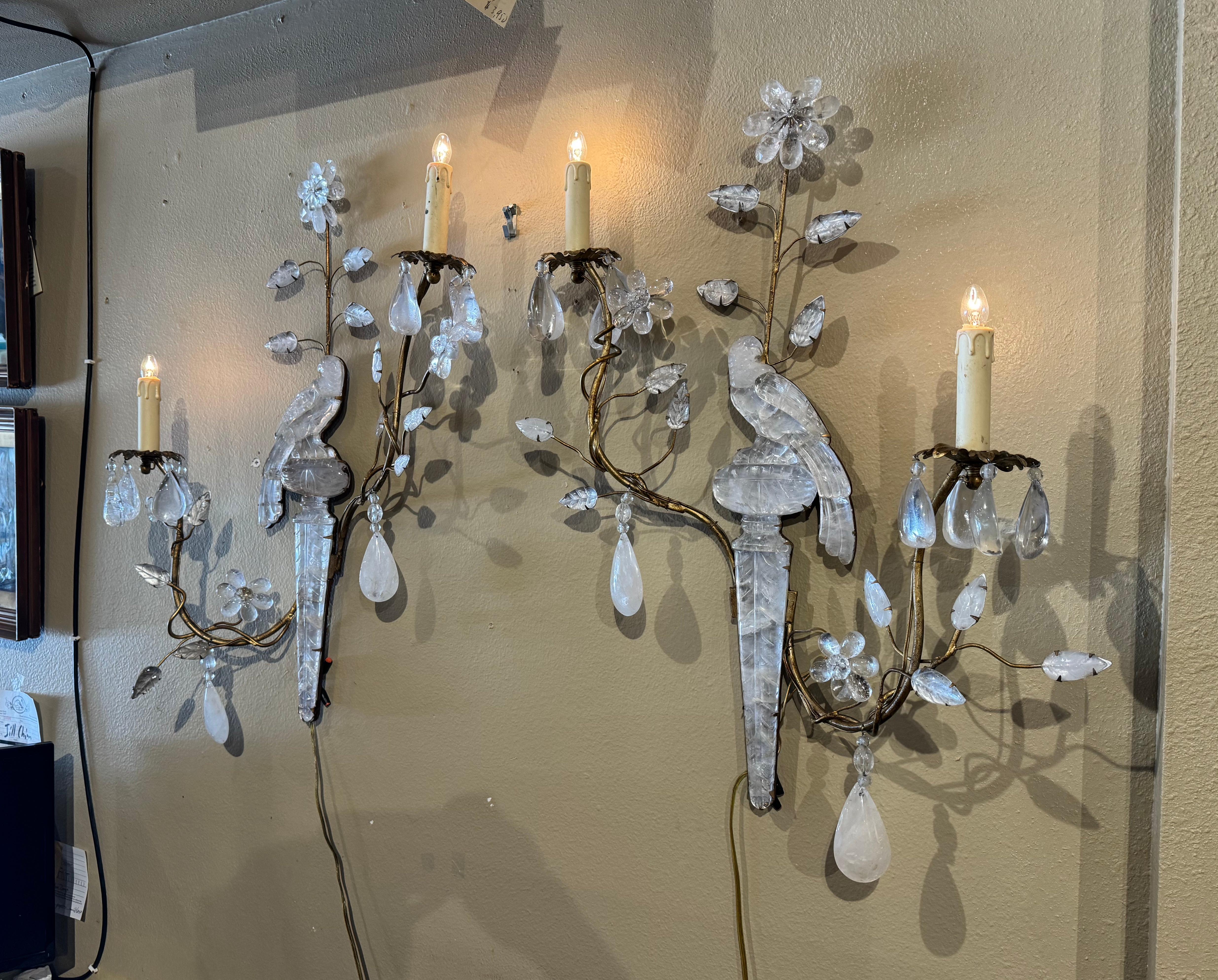 These elegant wall lights were created in France circa 1960. Attributed to Maison Bagues, each sconce with two metal arms is decorated with a rock crystal parrot perched on an urn with crystal form flowers and leaves throughout. Both fixtures have