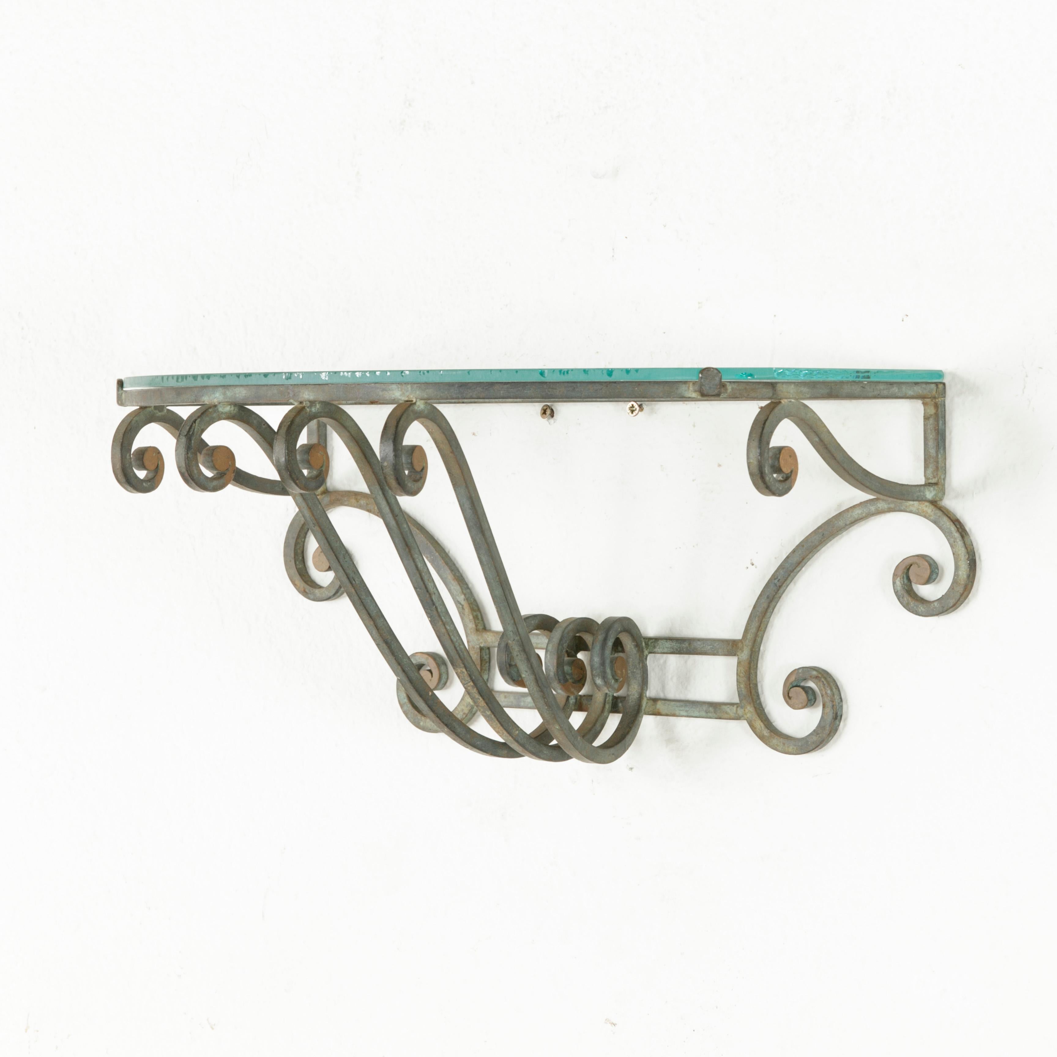 This pair of midcentury French iron wall sconces features handwrought scrolling ironwork and a verdigris patina with painted gold accents. A glass shelf rests on the demilune platform top of each bracket, providing display space. The glass may be