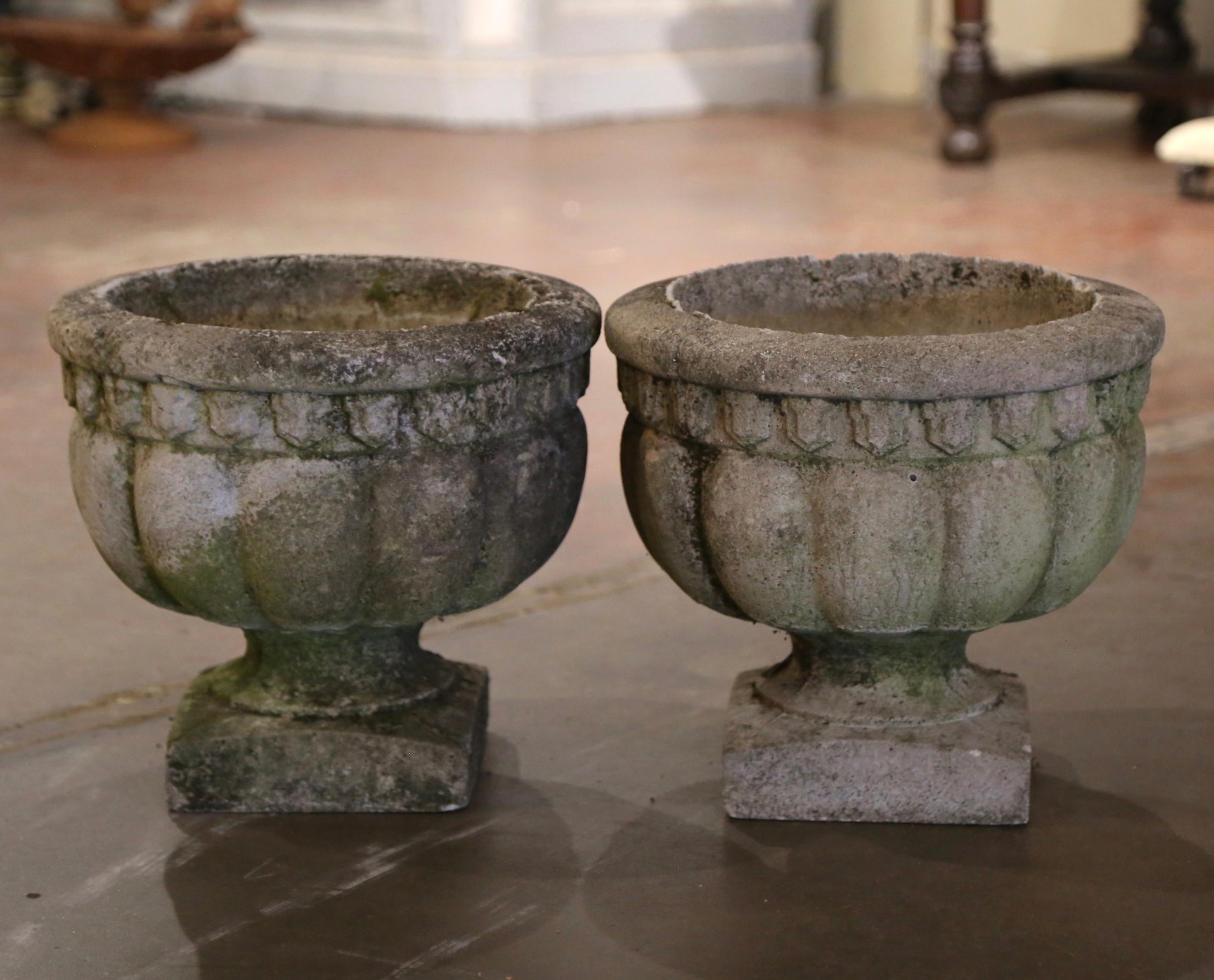 Crafted in France circa 1950 in the neoclassical style and made of cast stone, the pair of vintage stone planters have a stately, elegant style. Each vase stands on a square base over a gadrooned body with decorative architectural motifs around the