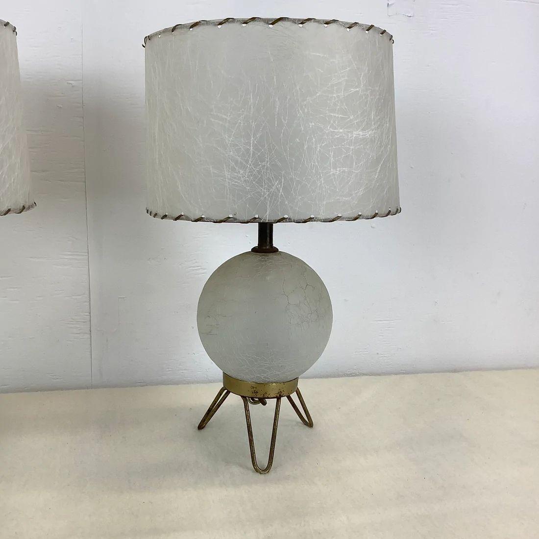 This striking pair of Mid-Century Modern table lamps feature unique crackled glass ball design set on brass hairpin legs and topped with unique skin style shades. The delicate matching pair make an impressive addition for their small Size, an