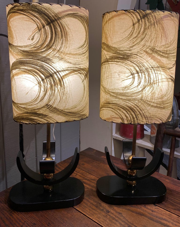 A fine pair of Mid Century Hollywood Regency table lamps with ebonized wood and brass bases, and original oval shades. The lamps are in very good overall condition, in good working order, with some brass finish wear, minor edge imperfections, and