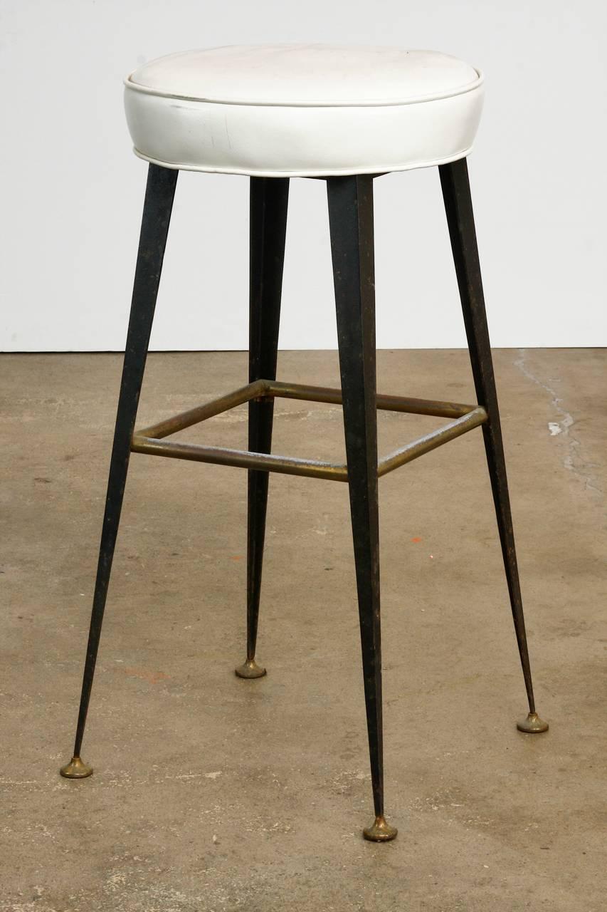 Interesting pair of midcentury industrial bar stools constructed from iron and metal and upholstered in white vinyl. Features tapered iron legs with contrasting brass metal feet and square stretchers. Evoking a steampunk style the hard industrial
