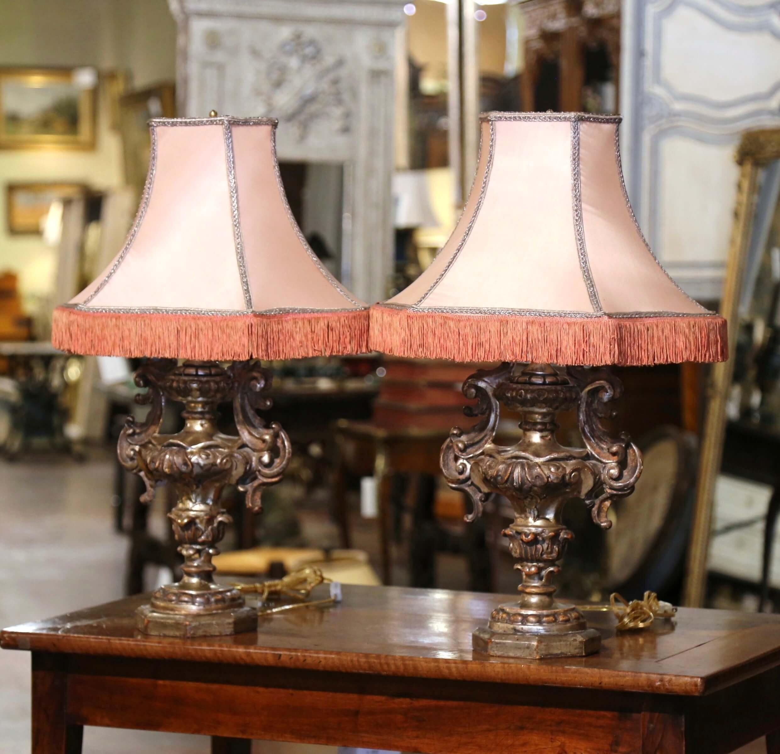 Place these elegant antique lamps on bedside tables or on top of a console in an entry! Crafted in Italy circa 1960, each urn formed lamp features intricately carved bases with scrollwork, floral motifs, and dressed with acanthus leaf handles. The