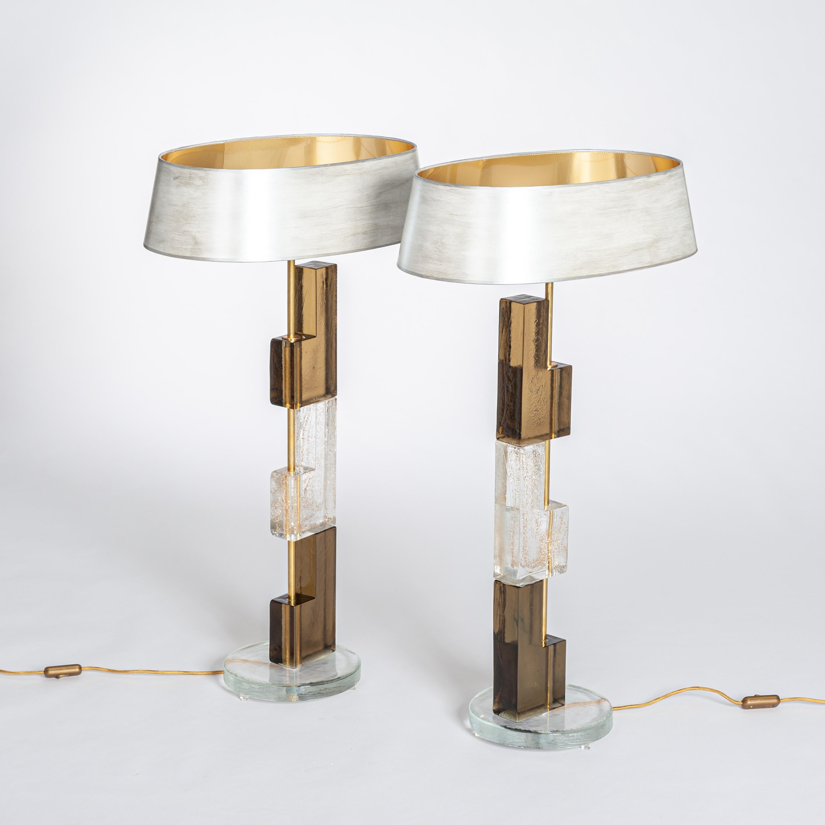 Pair of Murano glass table lamps made of rectangular elements in clear and amber-bronze-brown glass on a round base. 
The surface of the glass elements is slightly textured. The transparent element has gold particles in the glass mass.
Oval shades