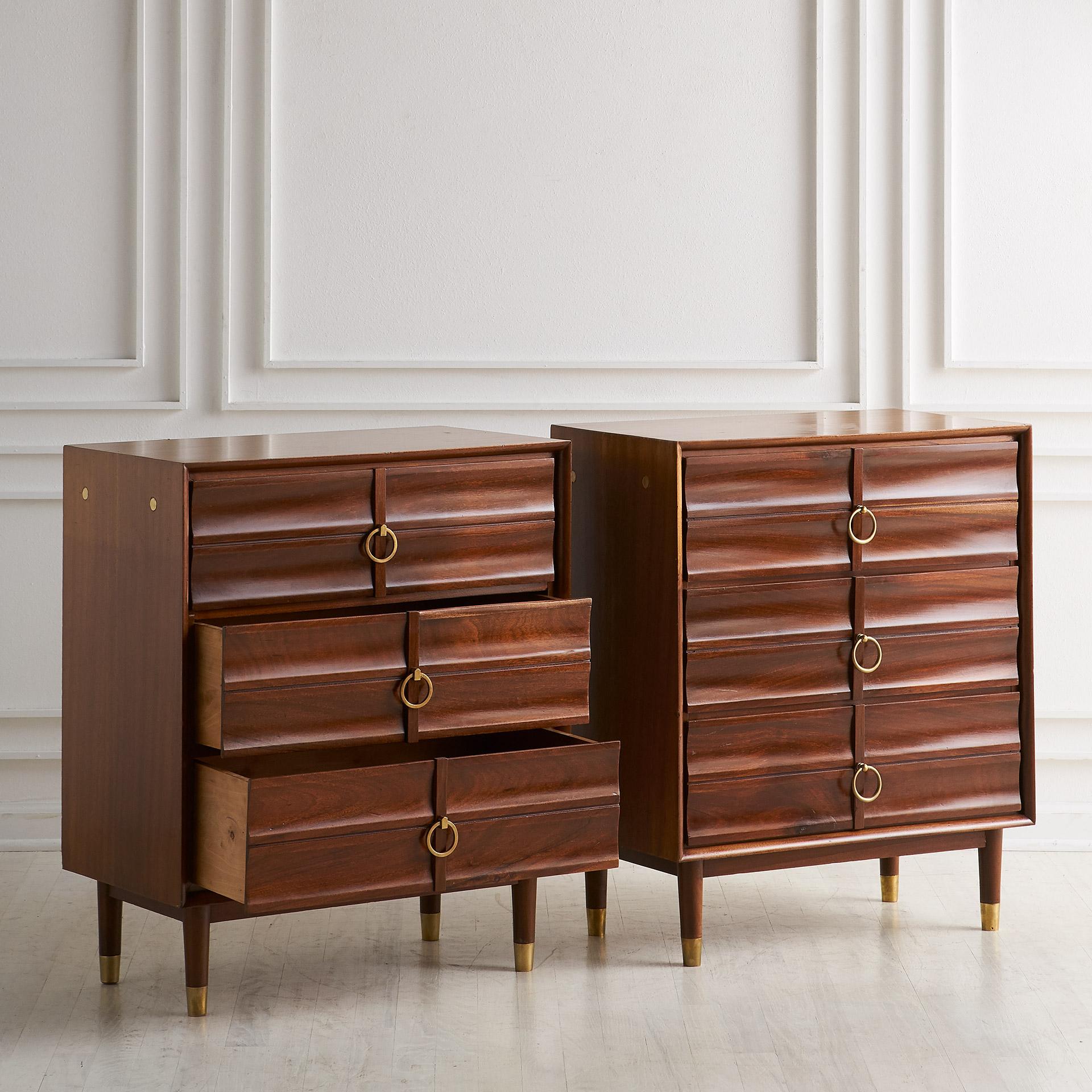 A pair of Mid-Century Modern three-drawer cabinets featuring curved drawer fronts with brass rings circa 1960. Nice inlaid brass detail on the sides and on the leg caps as well.