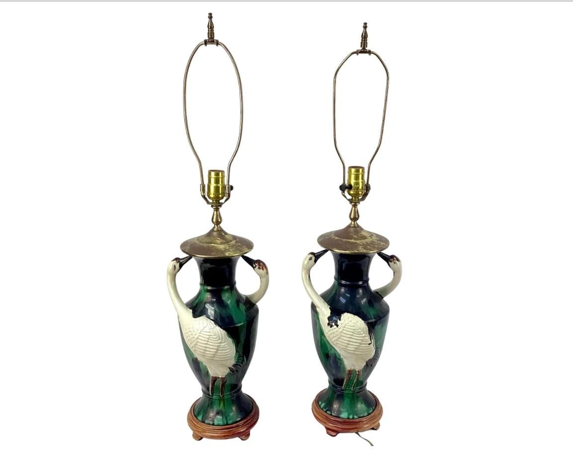 Beautiful pair of mid-century majolica vases fashioned into lamps. Each vase has shades of green and black with two white herons on either side with beaks touching neck of lamps. Vases are on wooden bases and have brass harps and finials. Height