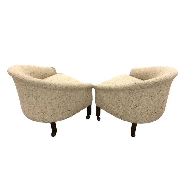 A pair of Mid Century Modern barrel back swivel chairs upholstered in neutral textured fabric.  Chairs are crafted in the style of Milo Baughman's chairs with curved back and arms.  The chairs sit on straight wood legs ending in casters.