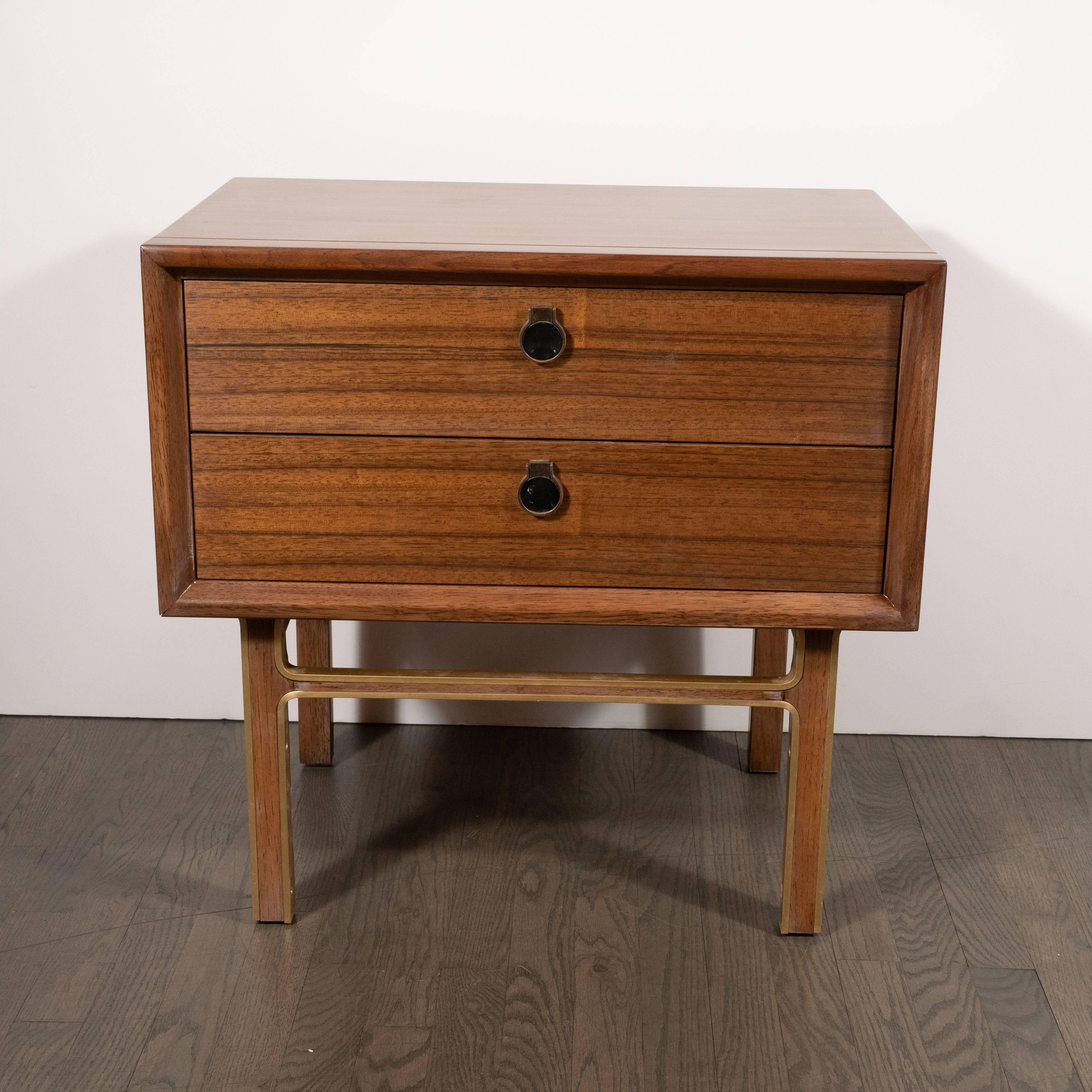 This refined and sophisticated pair of nightstands were realized in the United States, circa 1950. They feature rectangular bodies in book matched walnut with circular brass pulls. Additionally, they offer straight rectangular walnut legs wrapped in