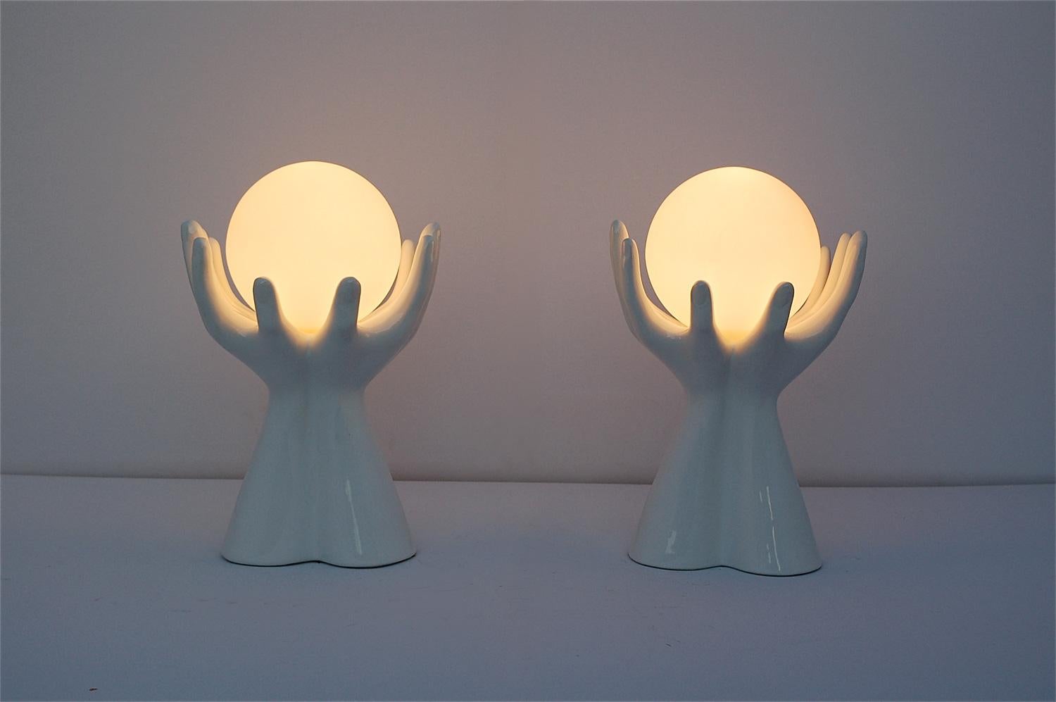 A matching pair of mid-20th century, 1960s white ceramic table lamps in the shape of a pair of hands clasping, holding a matte, opaque glass globe shade. The globe is held in place with a metal clasp. As a pair, they make an eclectic alternative to
