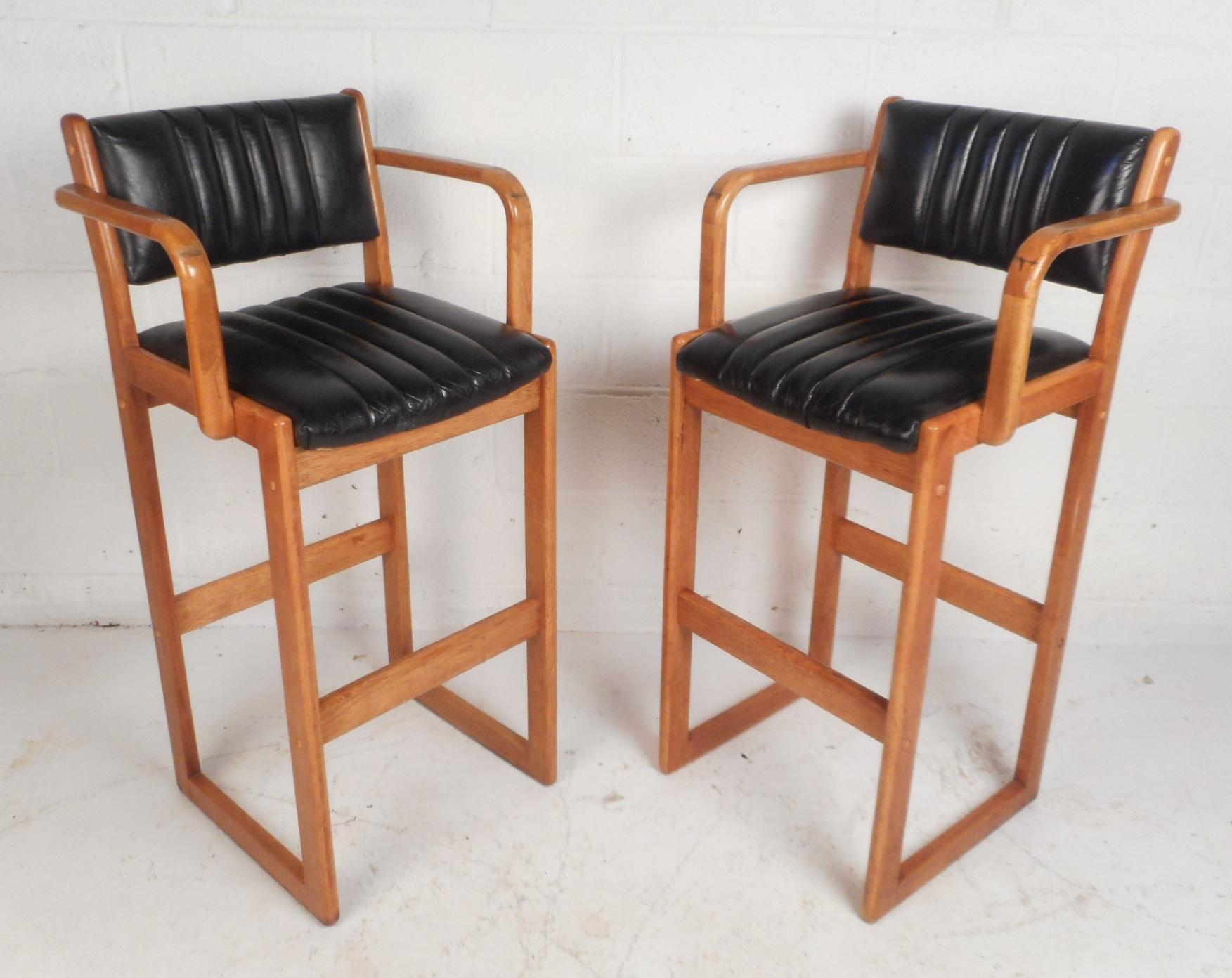 This beautiful pair of Danish modern bar stools feature a teak frame with sled legs. Unique design with high rounded arm rests, an angled backrest, and a black vinyl covered seat. This stunning pair offers plenty of comfort without sacrificing style