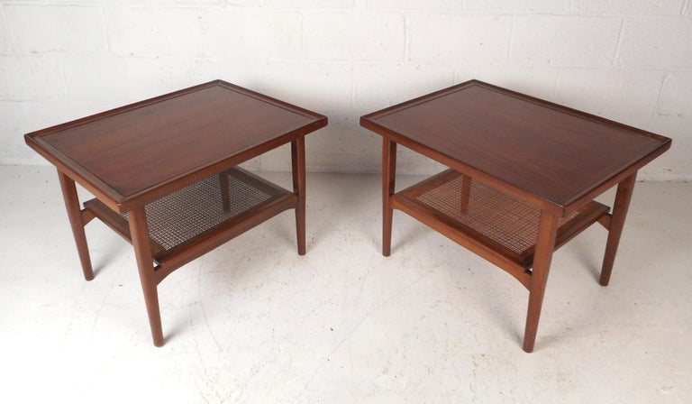 This gorgeous pair of vintage modern end tables feature a two-tier design with a lower woven cane shelf. Stylish design with raised edges along the top, a vintage dark walnut finish, and tapered legs. This beautiful pair of tables by Drexel make the