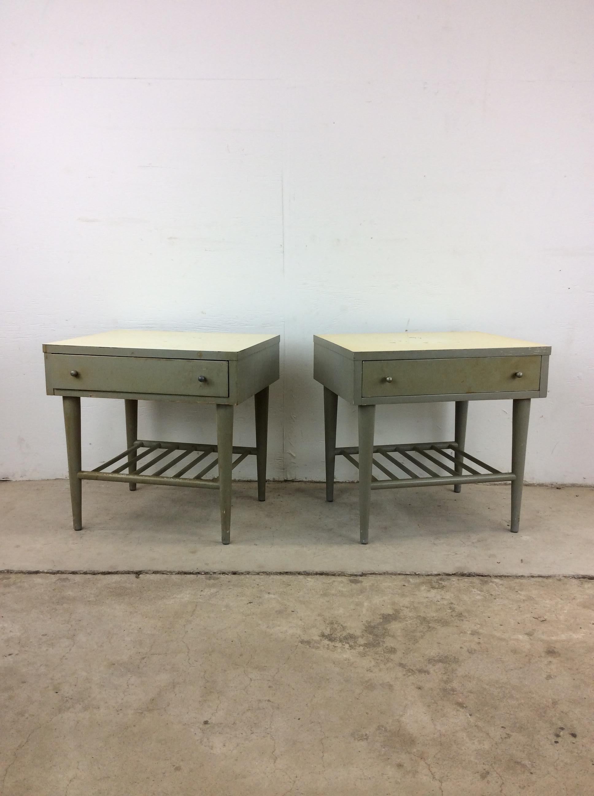This pair of mid century modern nightstands by American of Martinsville feature hardwood construction, gray painted finish, dovetailed drawer with brass accented hardware, slat open shelving below, white lacquer top, and tall tapered legs.