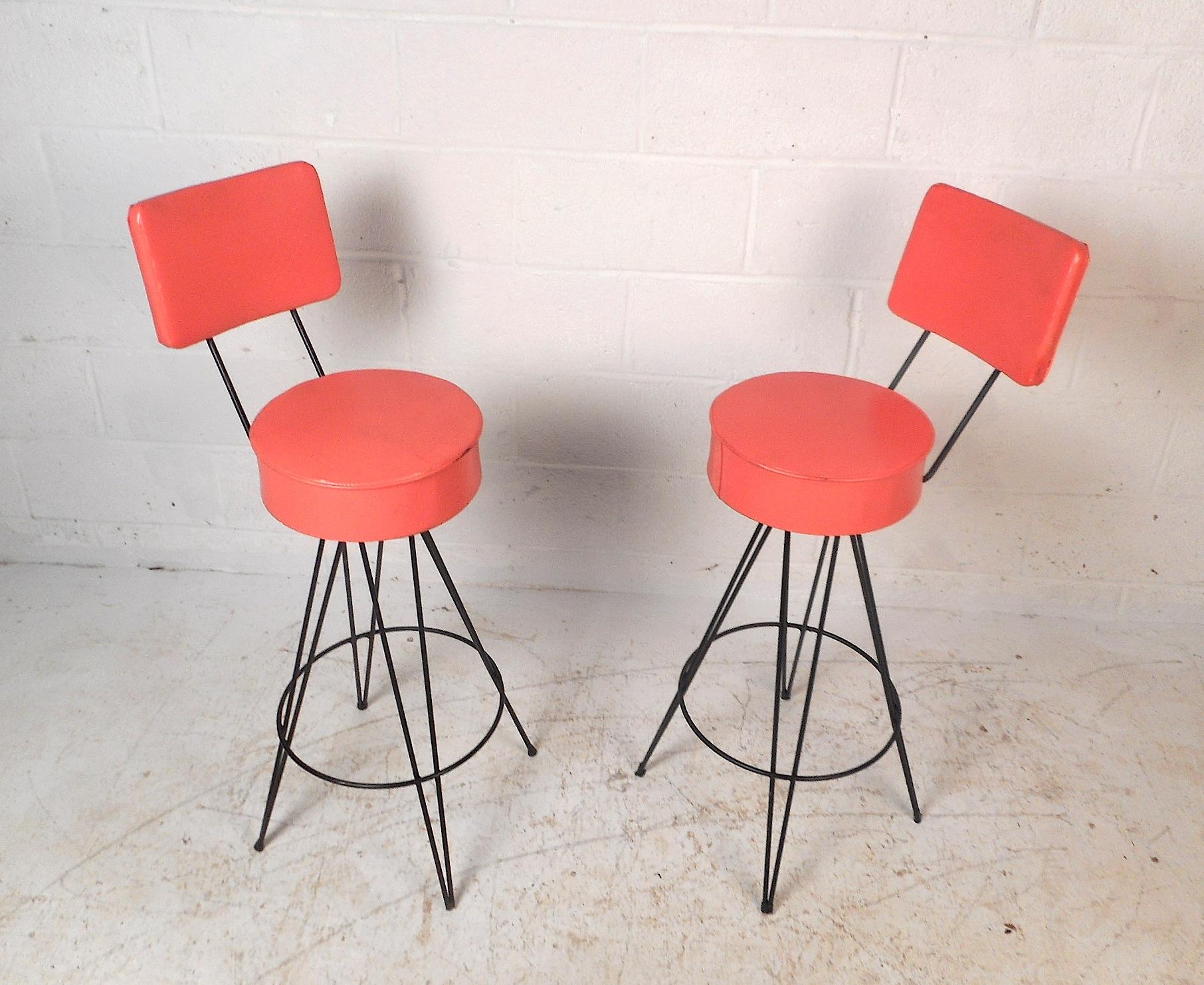 This stylish pair of vintage modern swivel bar stools feature a vibrant salmon colored vinyl upholstery along with sturdy wrought iron hairpin legs. The stools possess a thick seat cushion, padded backrests and a circular iron kickrest. These stools