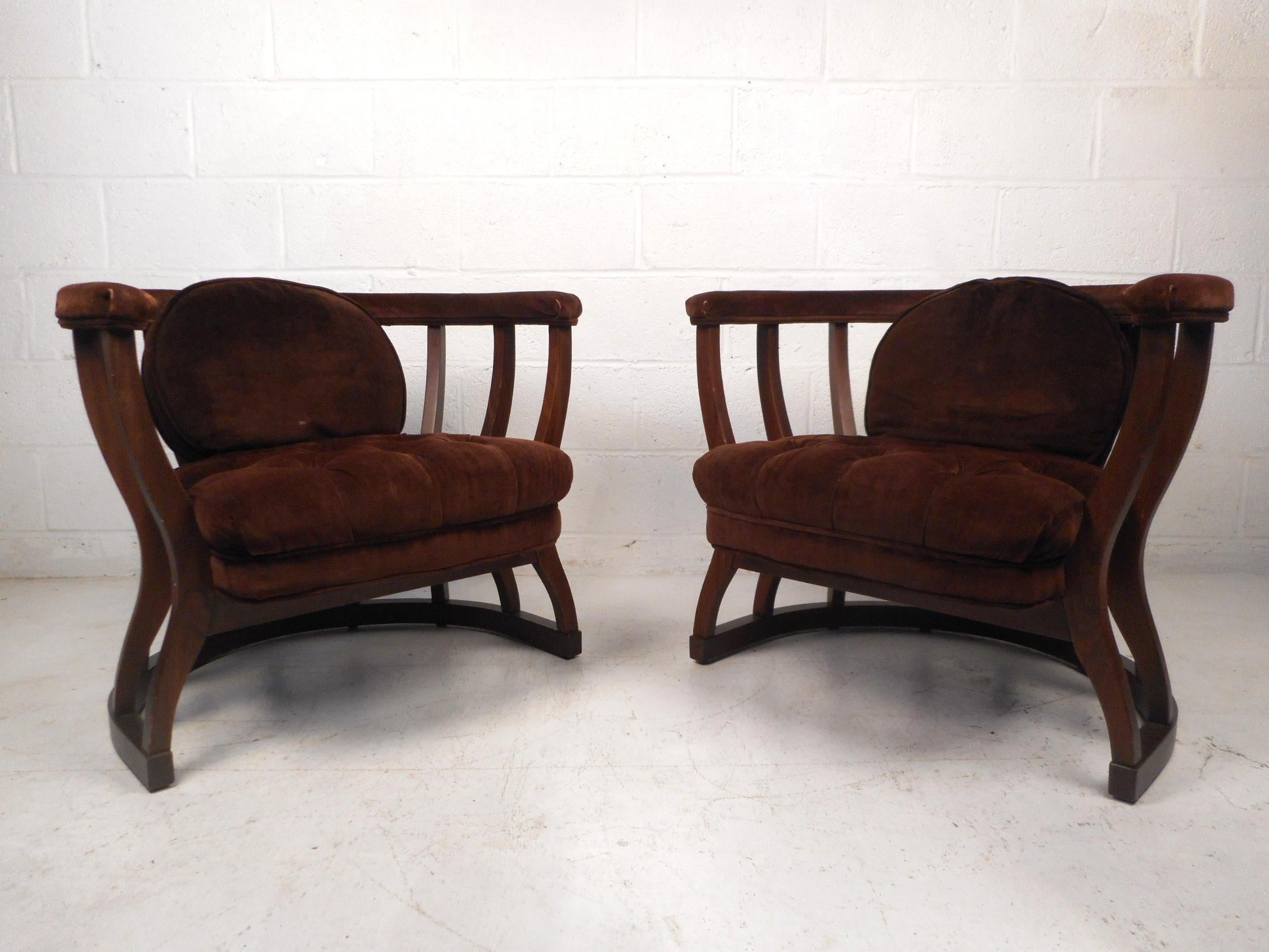 This impressive pair of Mid-Century Modern barrel back chairs feature a sturdy and sleek walnut frame with a dark finish, vintage brown tufted upholstery, removable backrest cushions, and padded armrests. The supports taper inward at seat level and