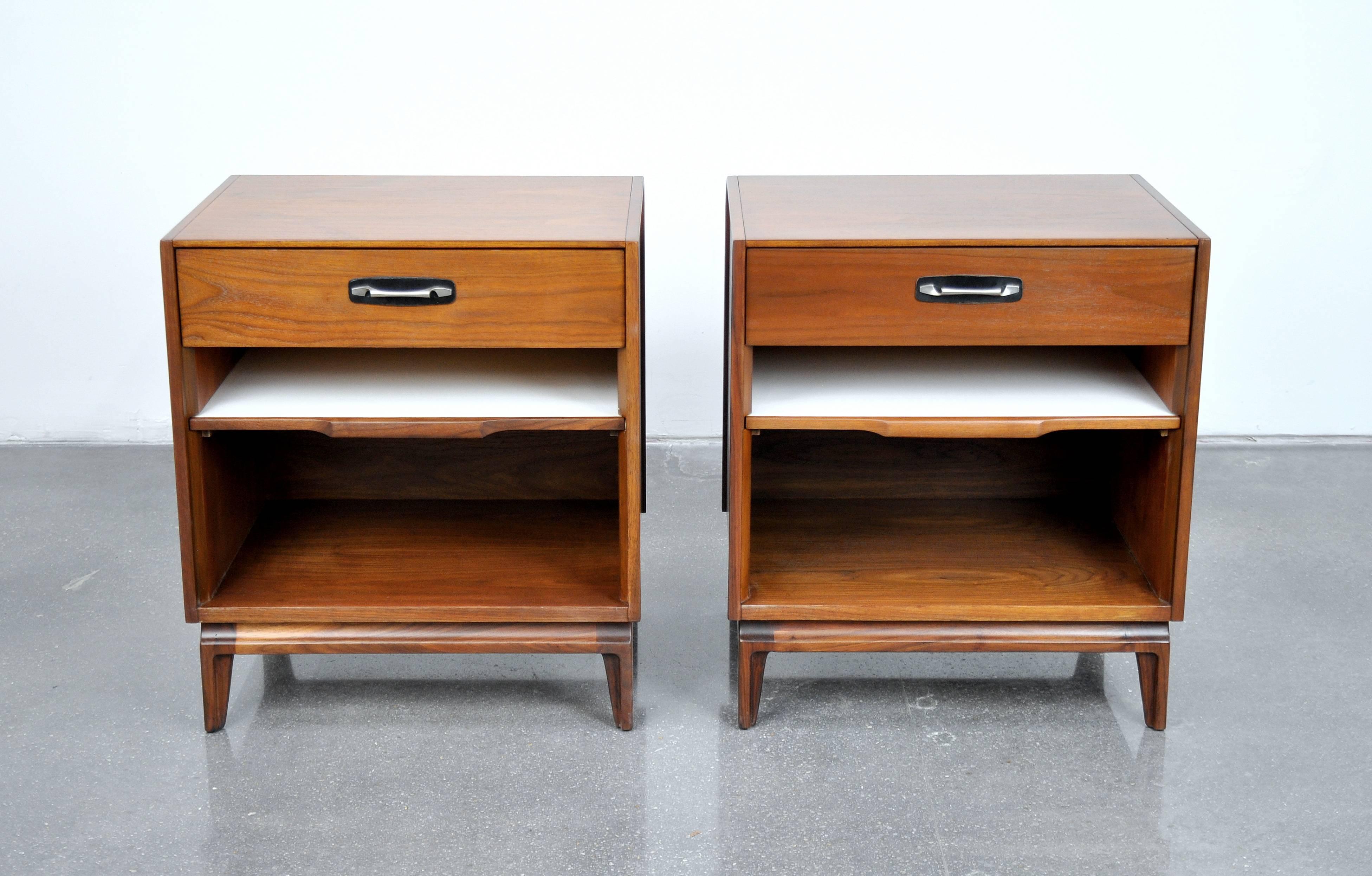 A vintage pair of Red Lion bedside tables, which could also be used as side or end tables, manufactured in the 1960s. Each table features a drawer with a black leather backed steel pull and a white slide out shelf, over an open cabinet space.
